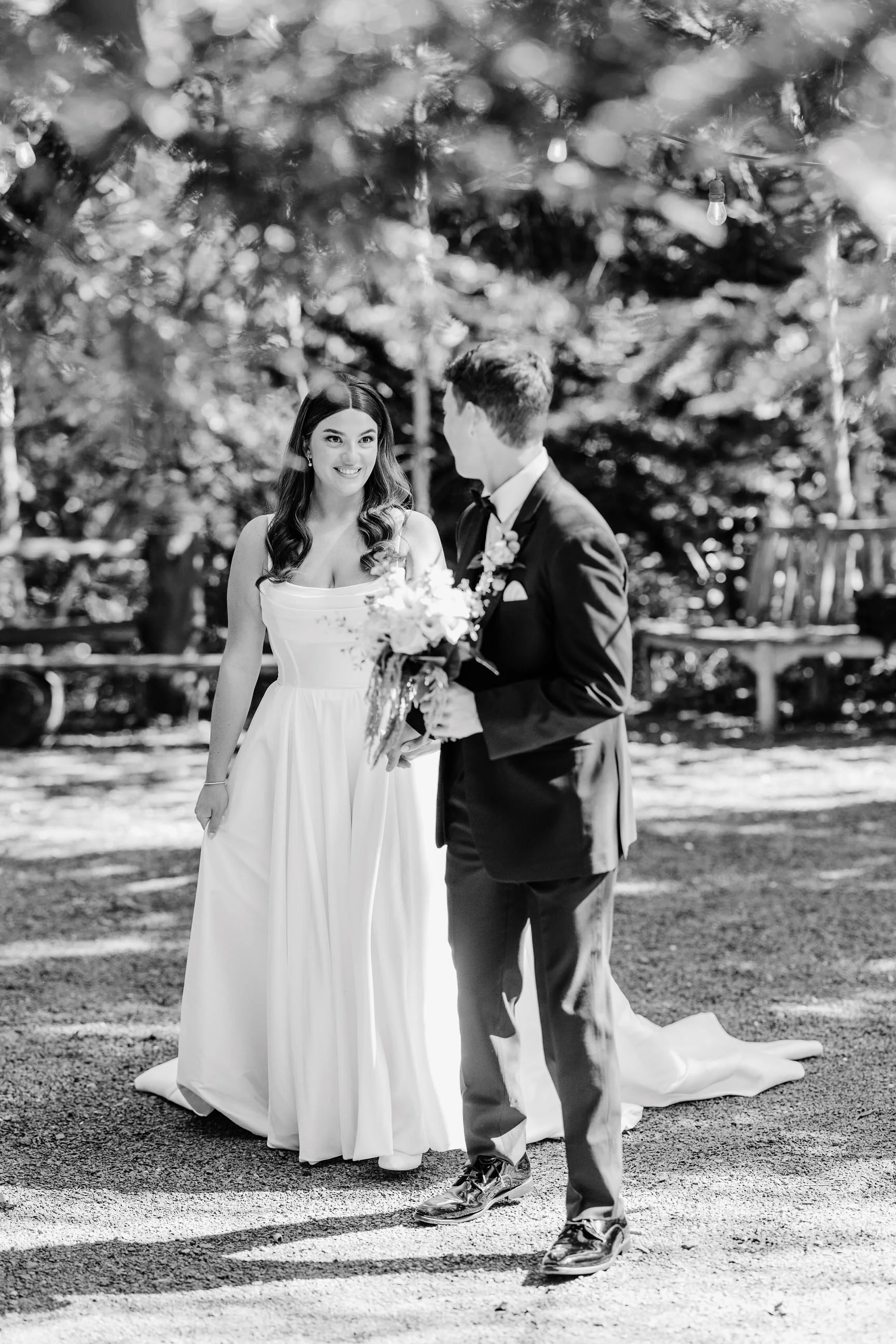 Black and white photograph of a bride and groom outdoors, with the groom offering a bouquet to the bride, surrounded by trees.