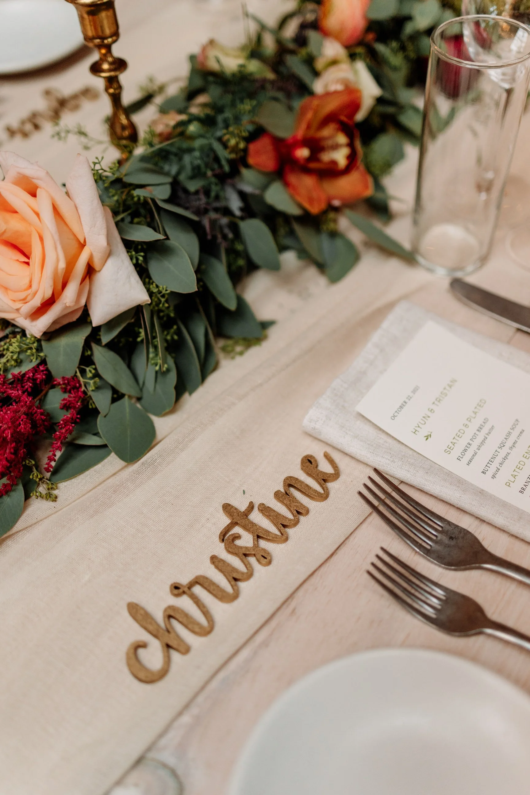 Wedding table setting with a floral centerpiece, a place card with the name 'Hyun & Travis', and a cloth napkin with the word 'Dessert' spelled in wooden letters. Silverware and a glass are also visible.