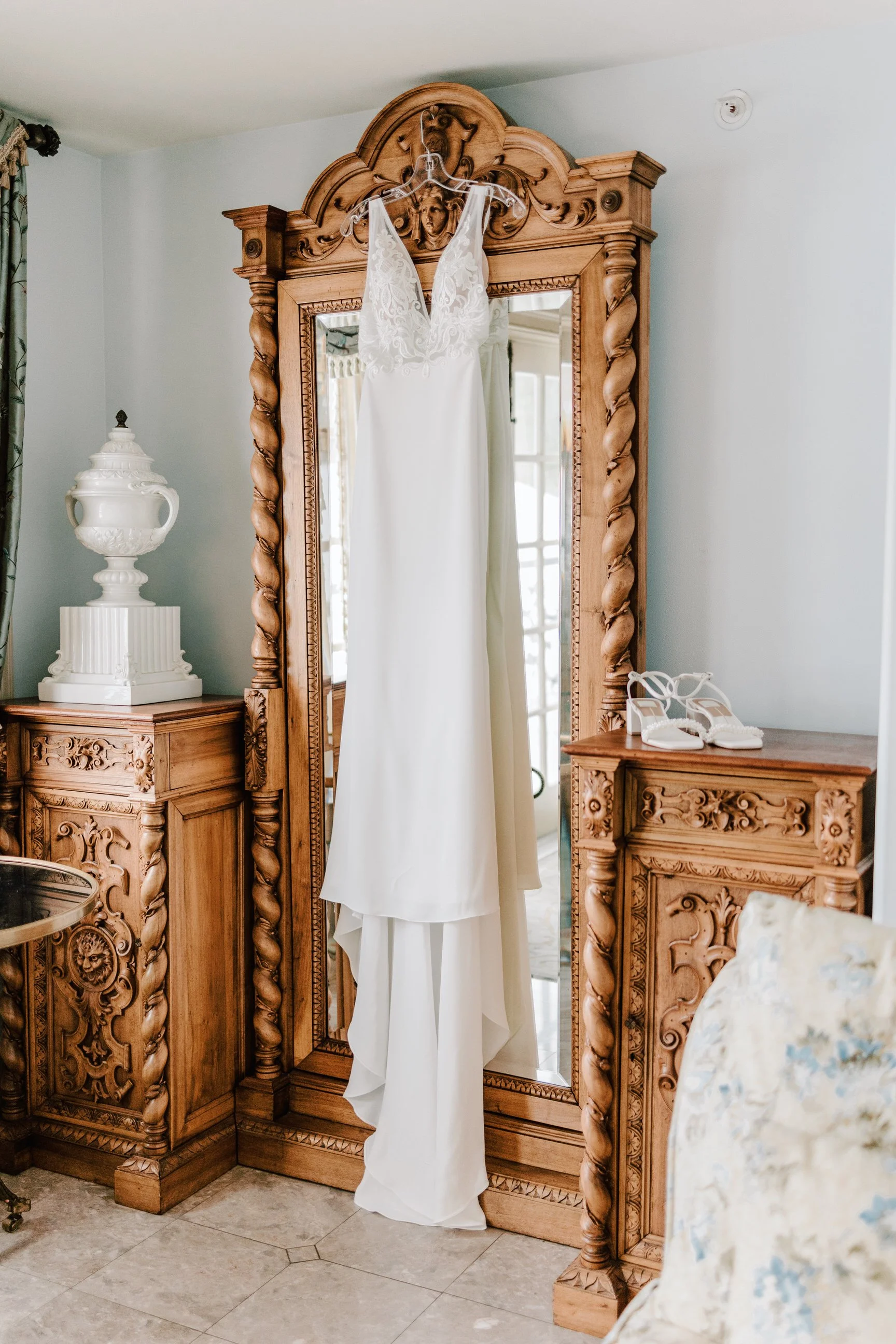 White wedding dress hanging on a decorative wooden mirror in a room with wooden furniture and a pair of white high-heeled shoes on a side table.
