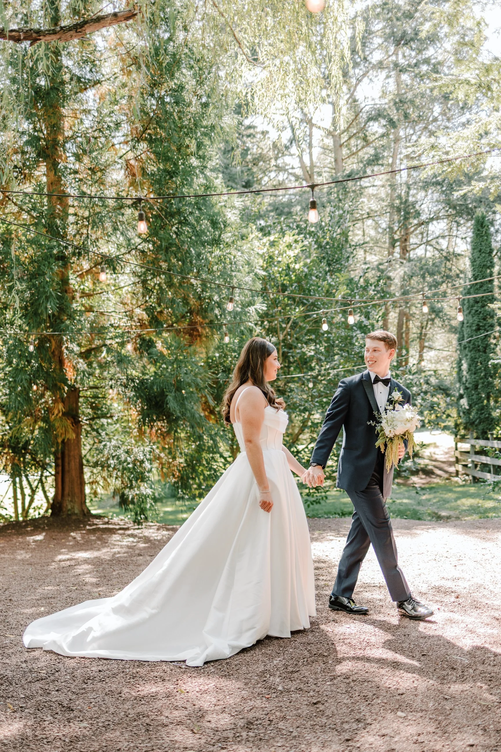 A bride and groom holding hands outdoors, surrounded by trees and string lights, during their wedding ceremony.