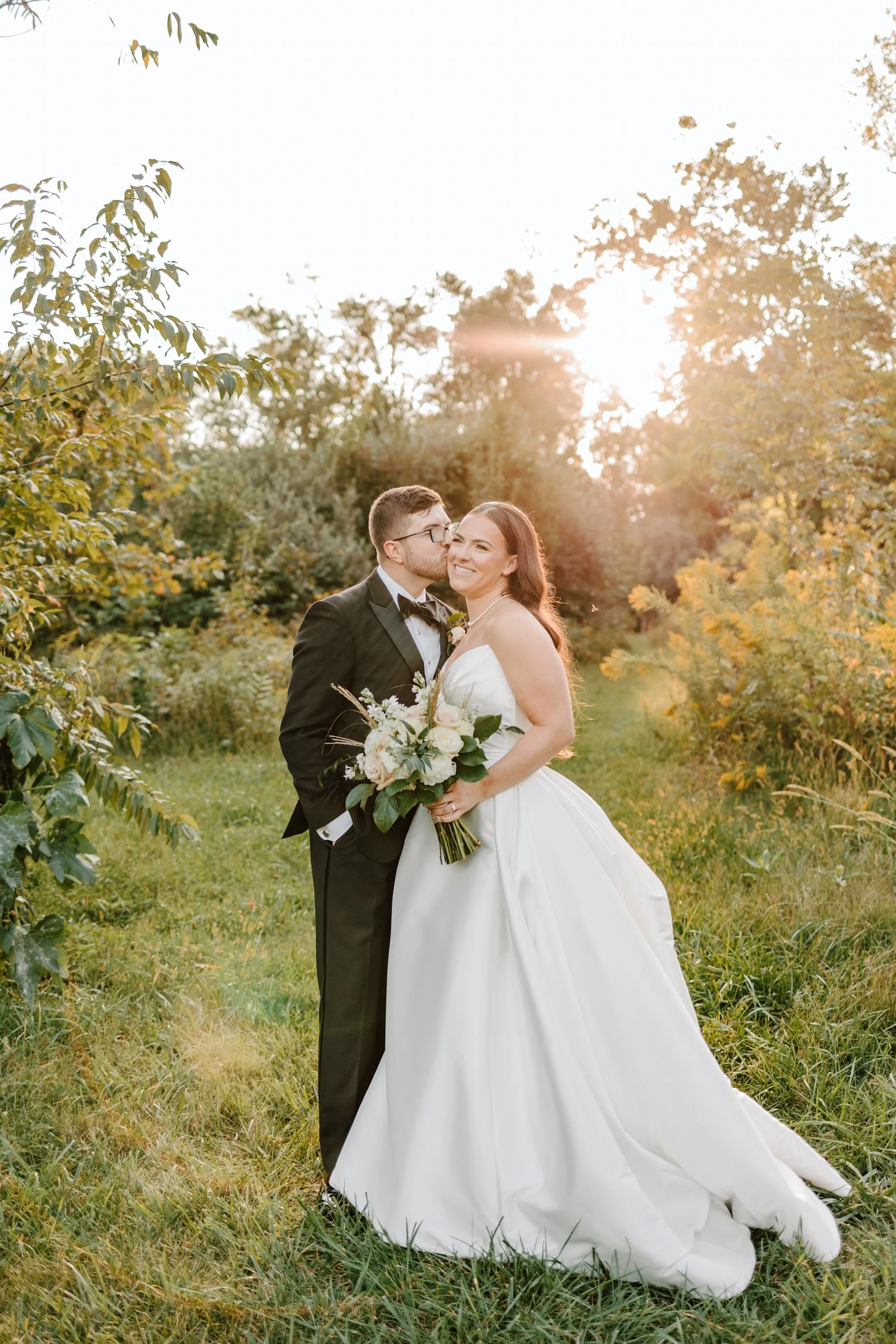 A newlywed couple stands outdoors on a grassy path, with the groom kissing the bride on the cheek. The bride, in a white wedding gown, is holding a bouquet of flowers and smiling. The groom is in a black tuxedo with a bow tie. The sun is setting behi