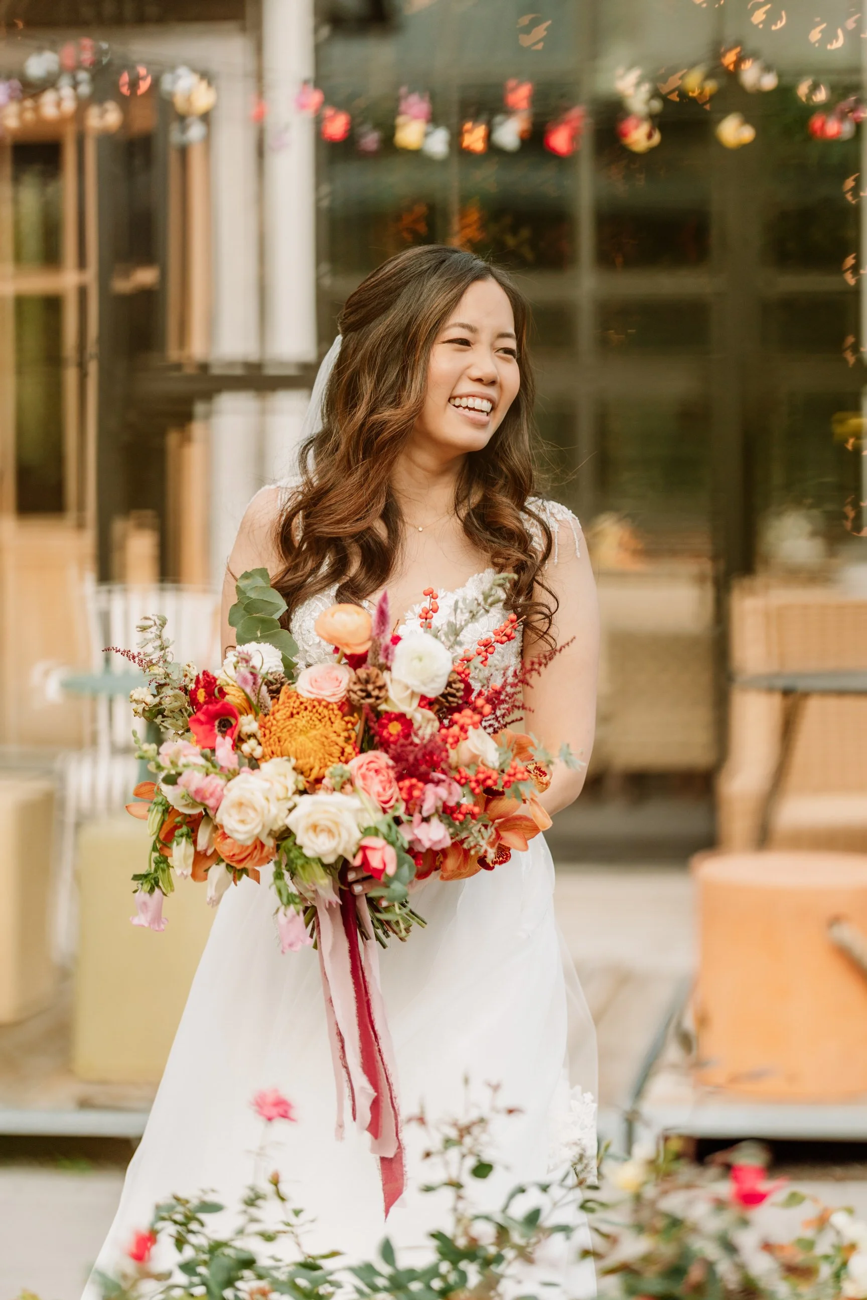 A happy woman in a white dress holding a large colorful bouquet of flowers, standing outdoors with blurred background of a glass building and festive string lights.