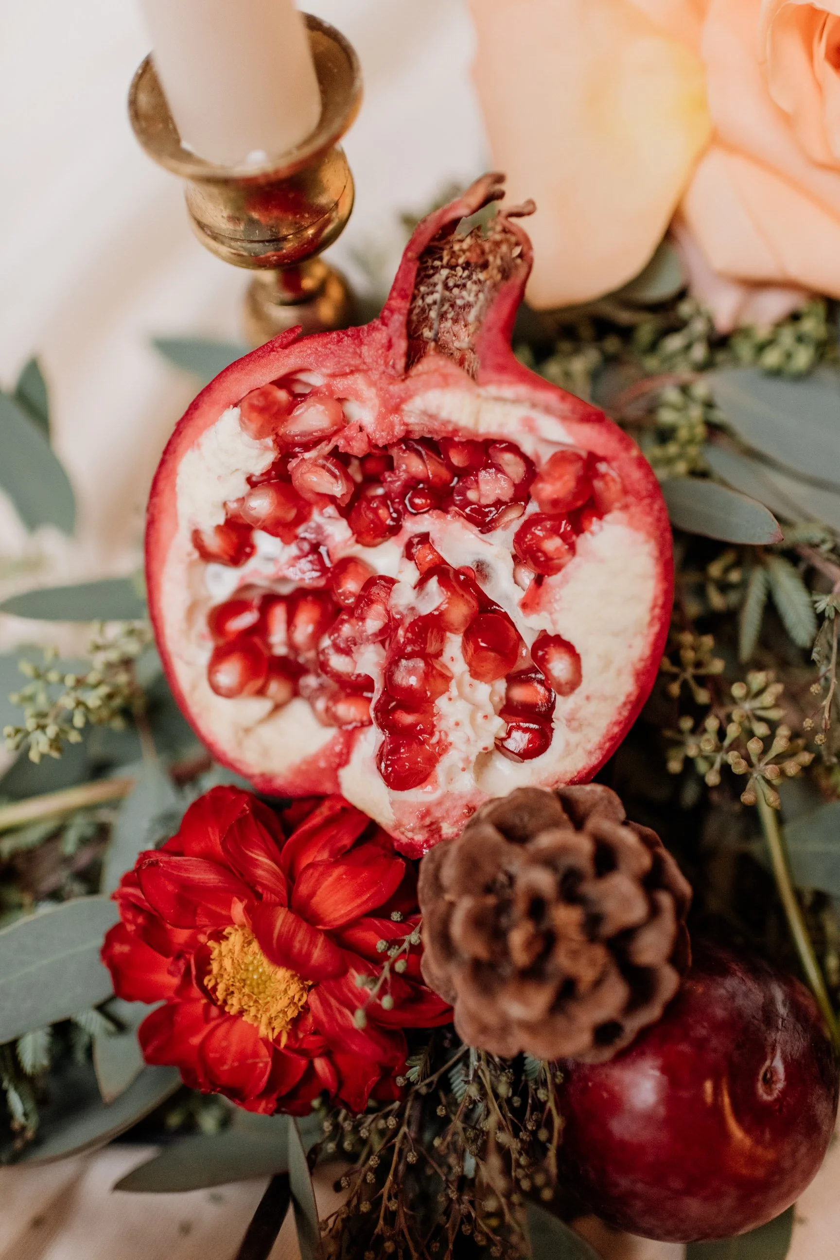 A halved pomegranate surrounded by a red flower, a pine cone, an apple, and greenery with a gold candle holder and a peach-colored rose in the background.