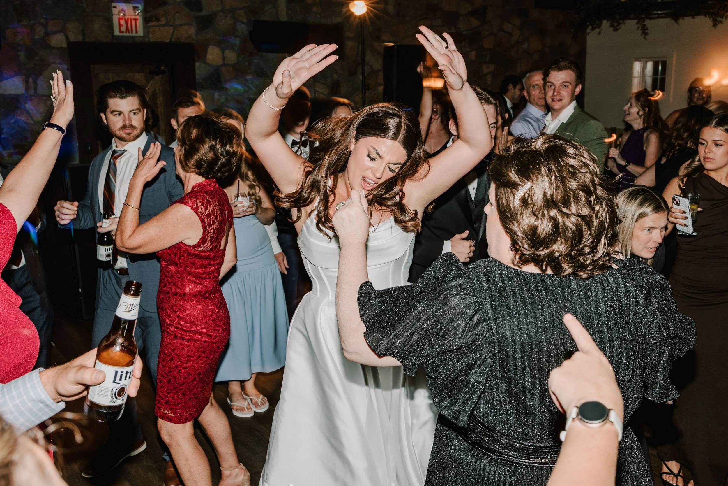 People dancing and celebrating at a party or wedding reception with a stone wall background. A woman in a white dress is dancing with a woman in a black dress, both enjoying the moment. Others are holding drinks and taking photos.