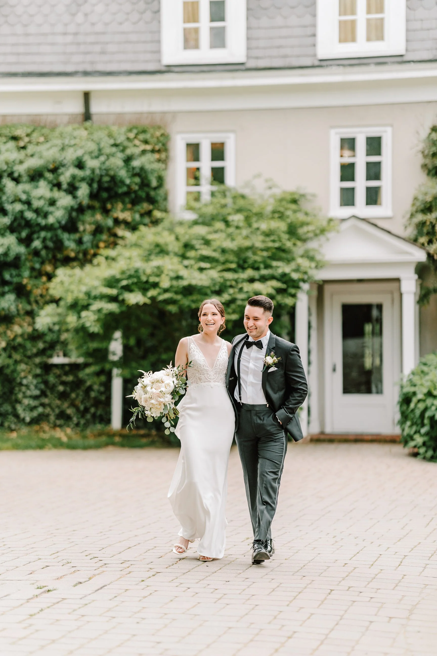 A bride and groom walking outdoors on a brick pathway, smiling, with a house and greenery in the background.