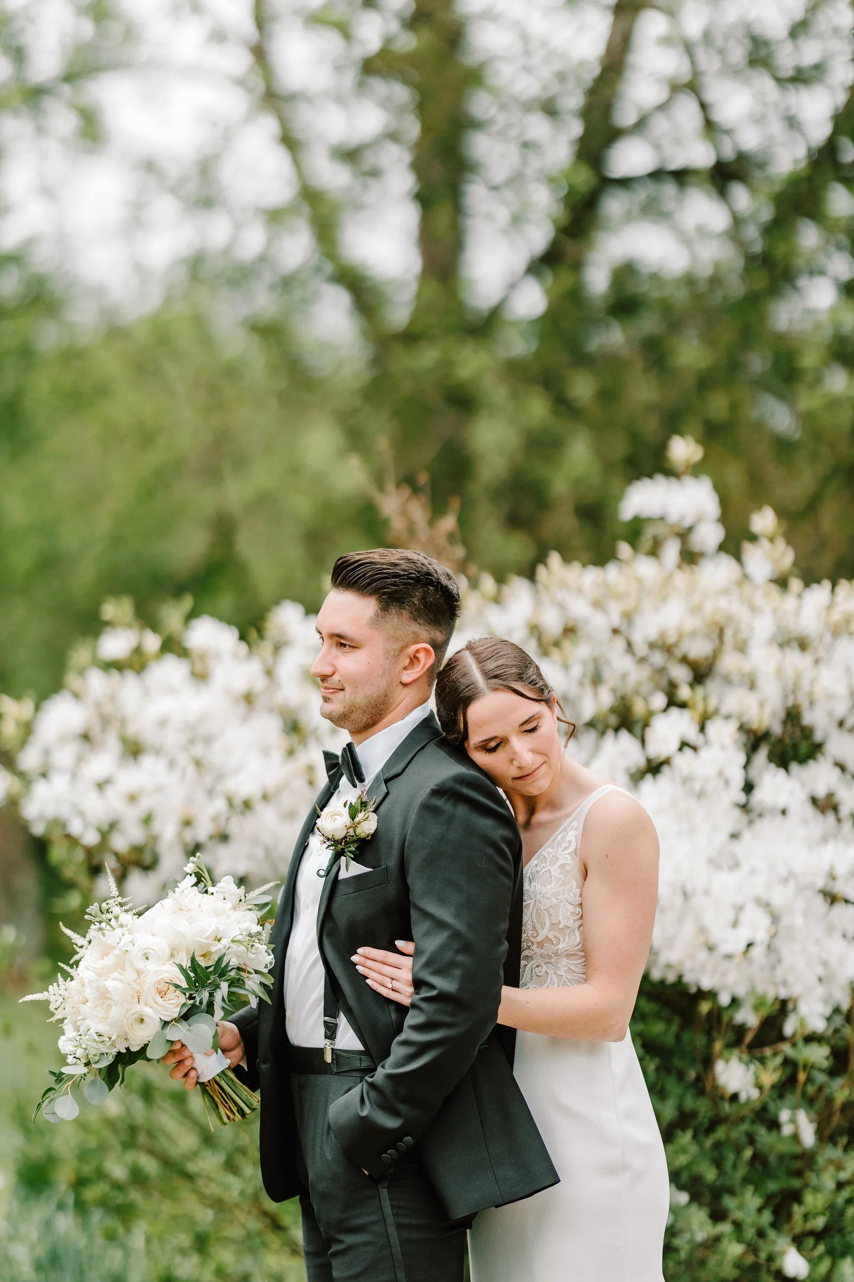 A bride and groom stand together outdoors, the bride resting her head on the groom's back, with white flowers in the background.