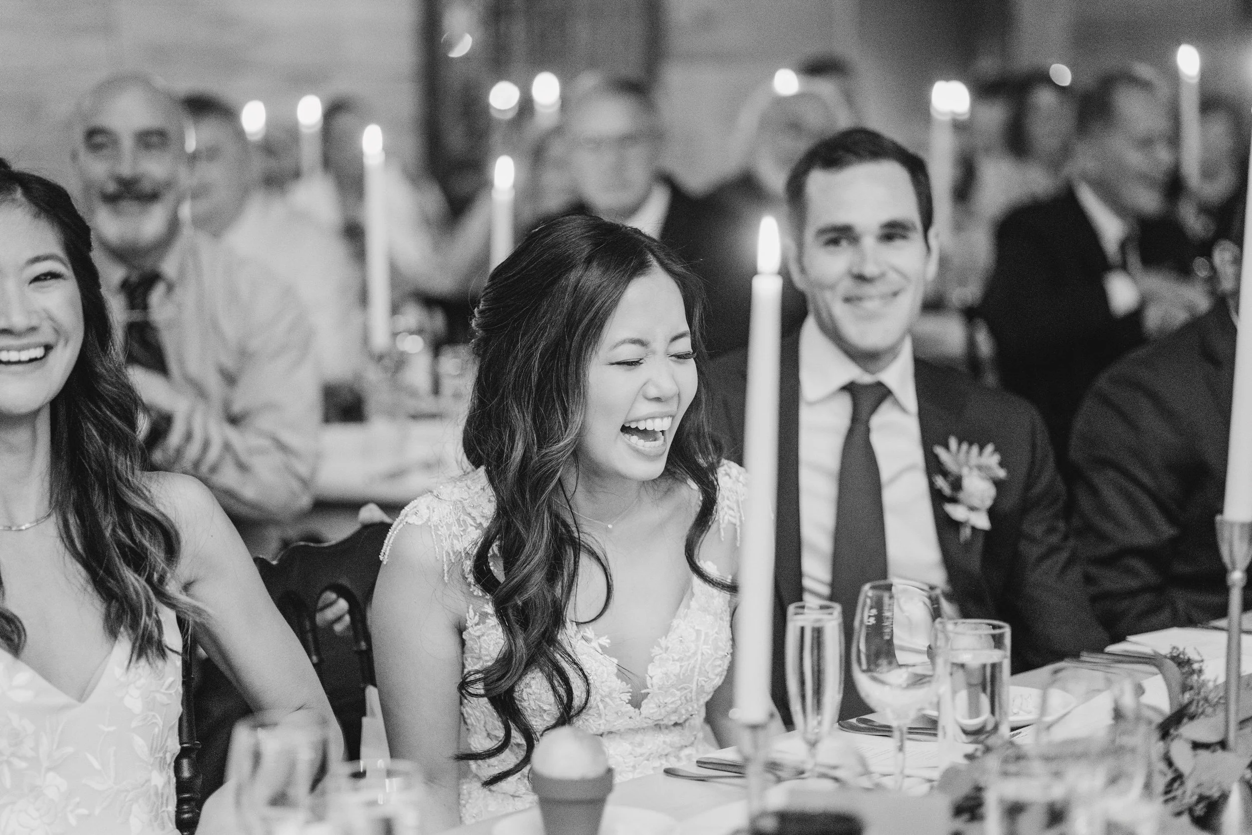 People enjoying a wedding reception, with a woman laughing and a man smiling at a table decorated with candles and glassware.