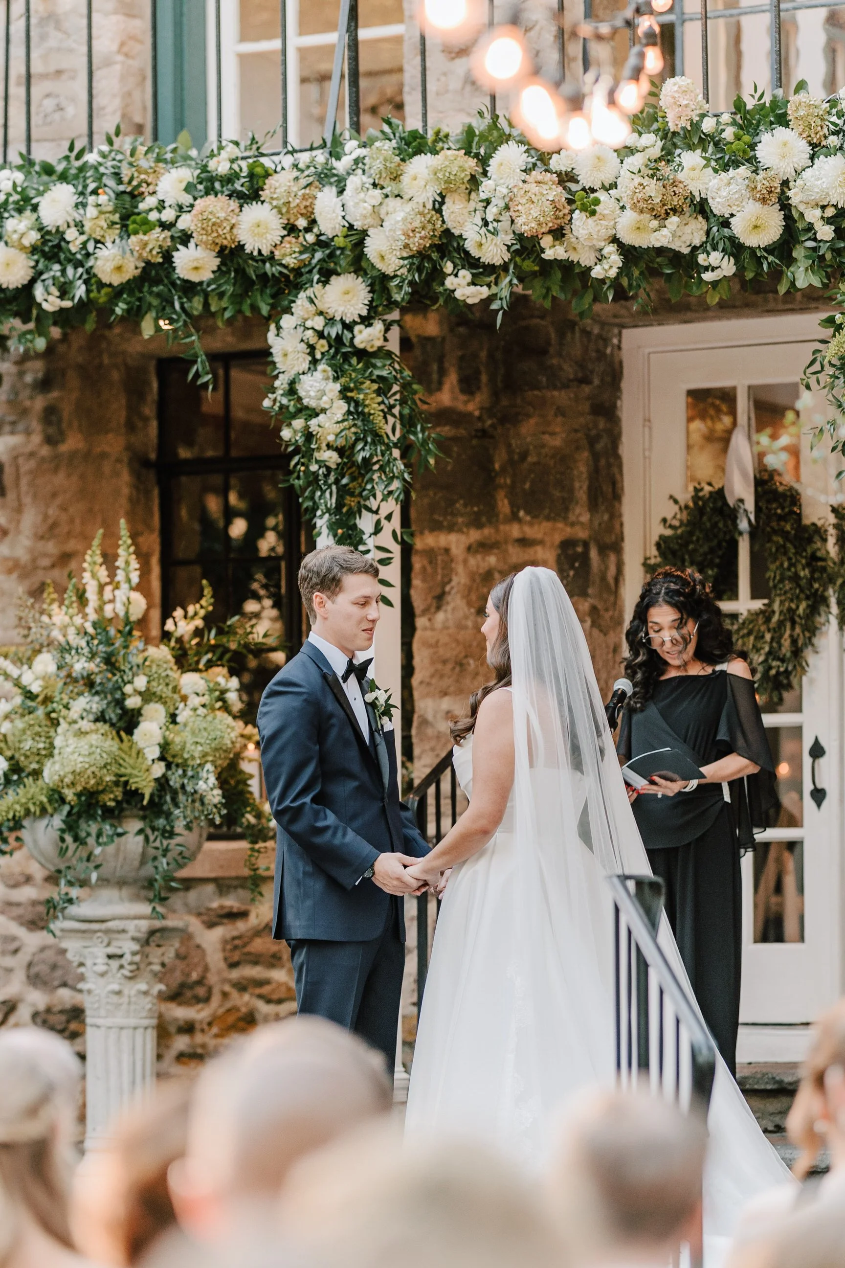 A wedding ceremony with a bride and groom standing hand in hand under a floral arch, with an officiant reading from a book, inside a rustic venue with stone walls and large windows.
