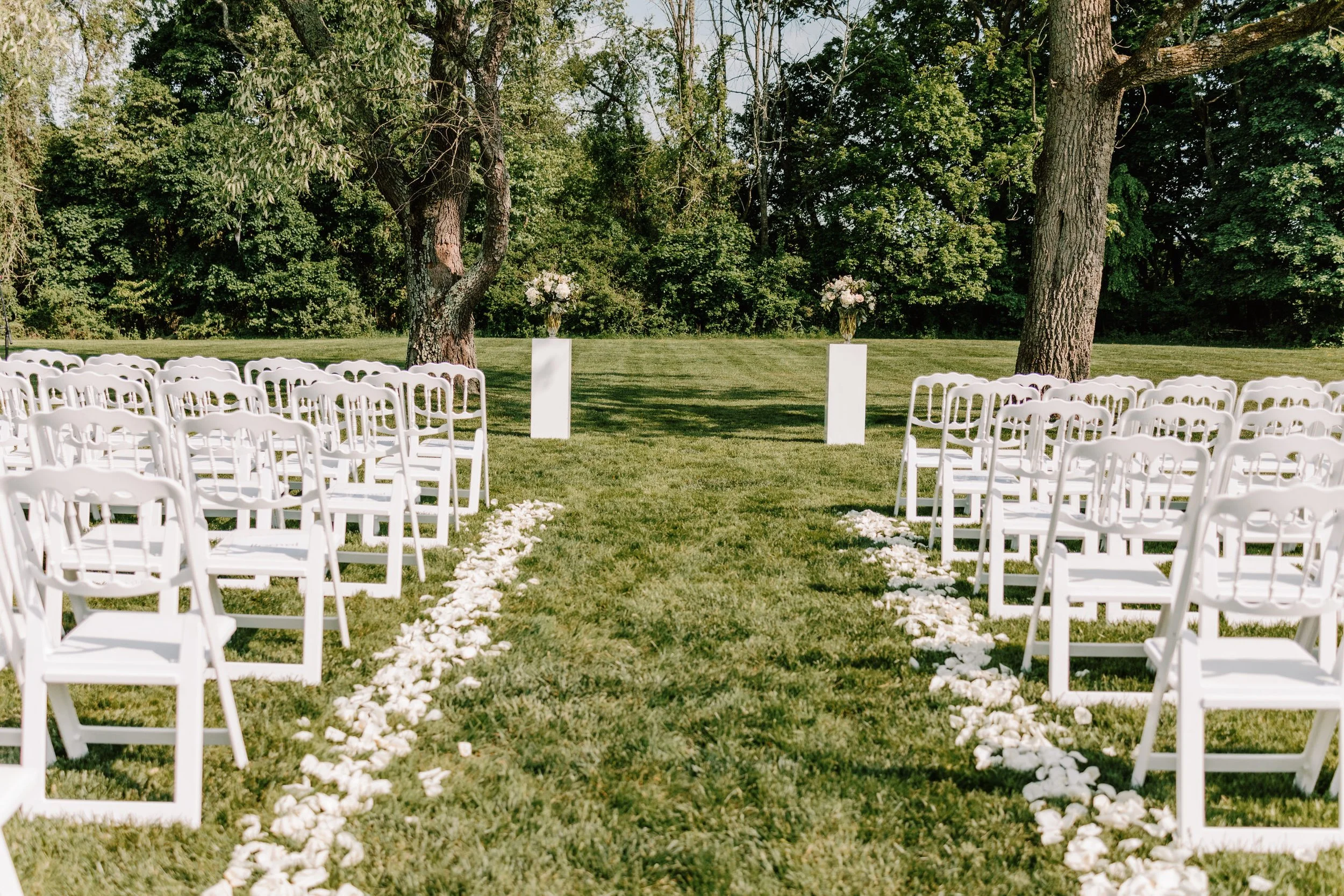 Outdoor wedding ceremony setup with white chairs arranged on a grassy area, flower petals scattered along the aisle, two white pedestals with floral arrangements, trees and greenery in the background.