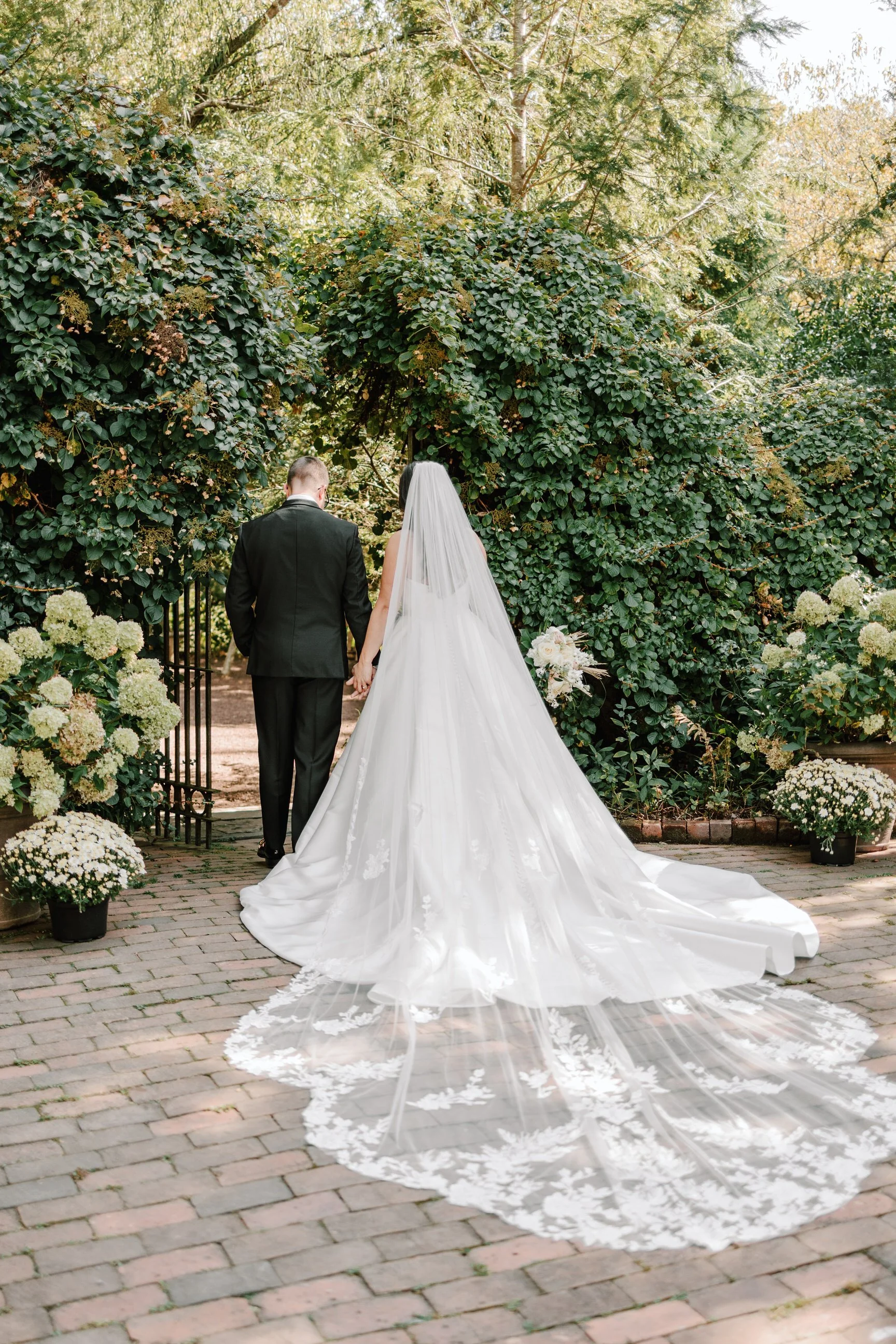 A bride and groom walking hand in hand through an outdoor garden archway, surrounded by greenery and white flowers during the daytime.