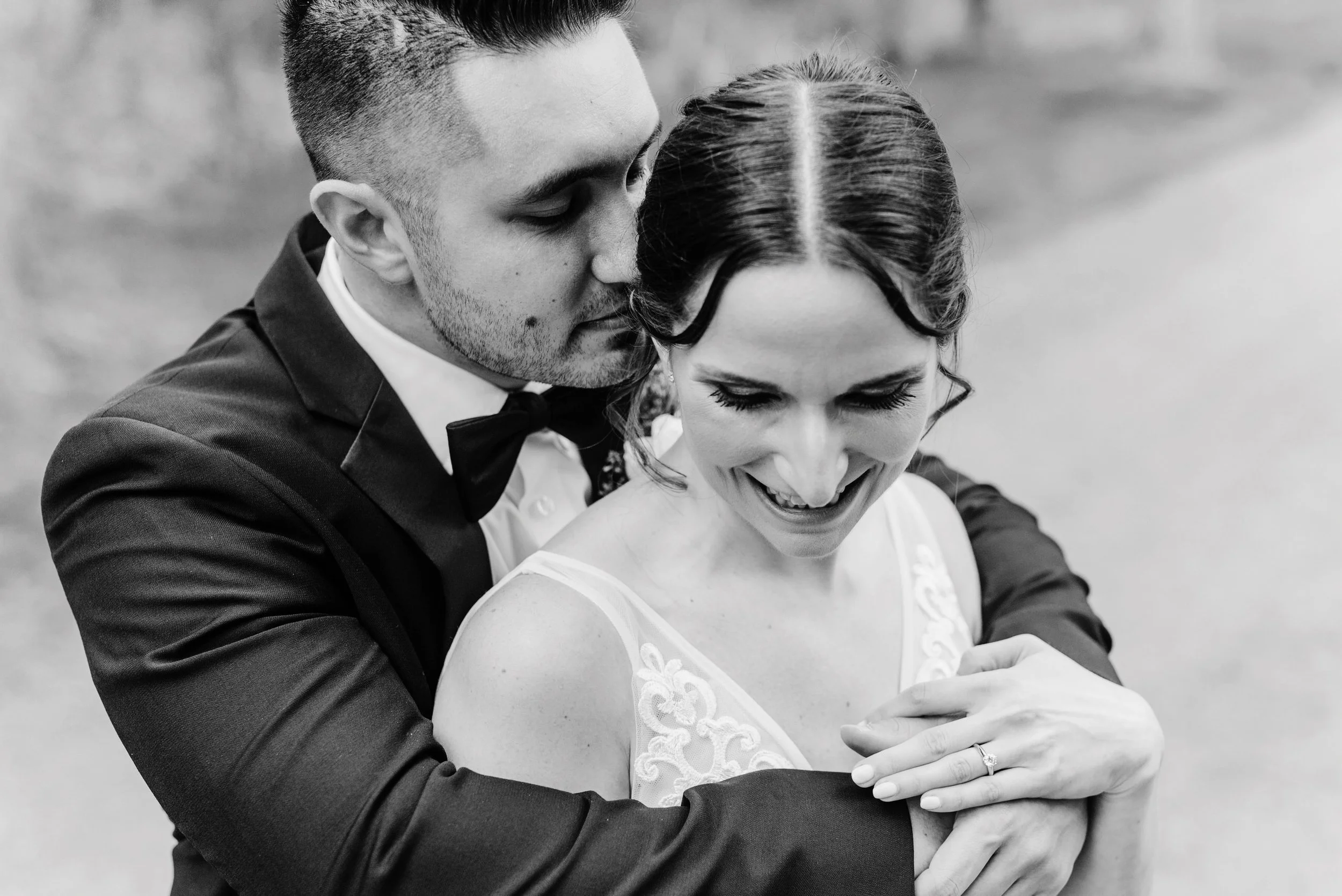 A black and white photo of a happy couple, with the man embracing the woman from behind, both smiling. The woman shows her engagement ring.
