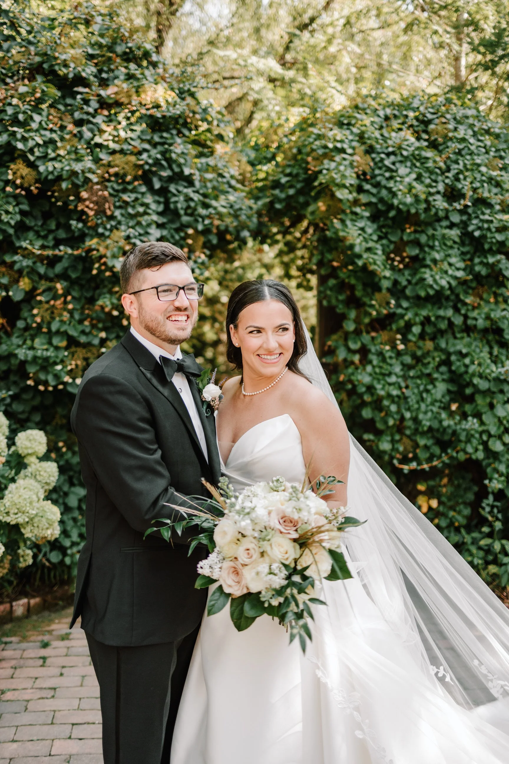 A newlywed couple in wedding attire smiling outdoors, surrounded by lush greenery, with the bride holding a bouquet of white and blush roses and greenery.