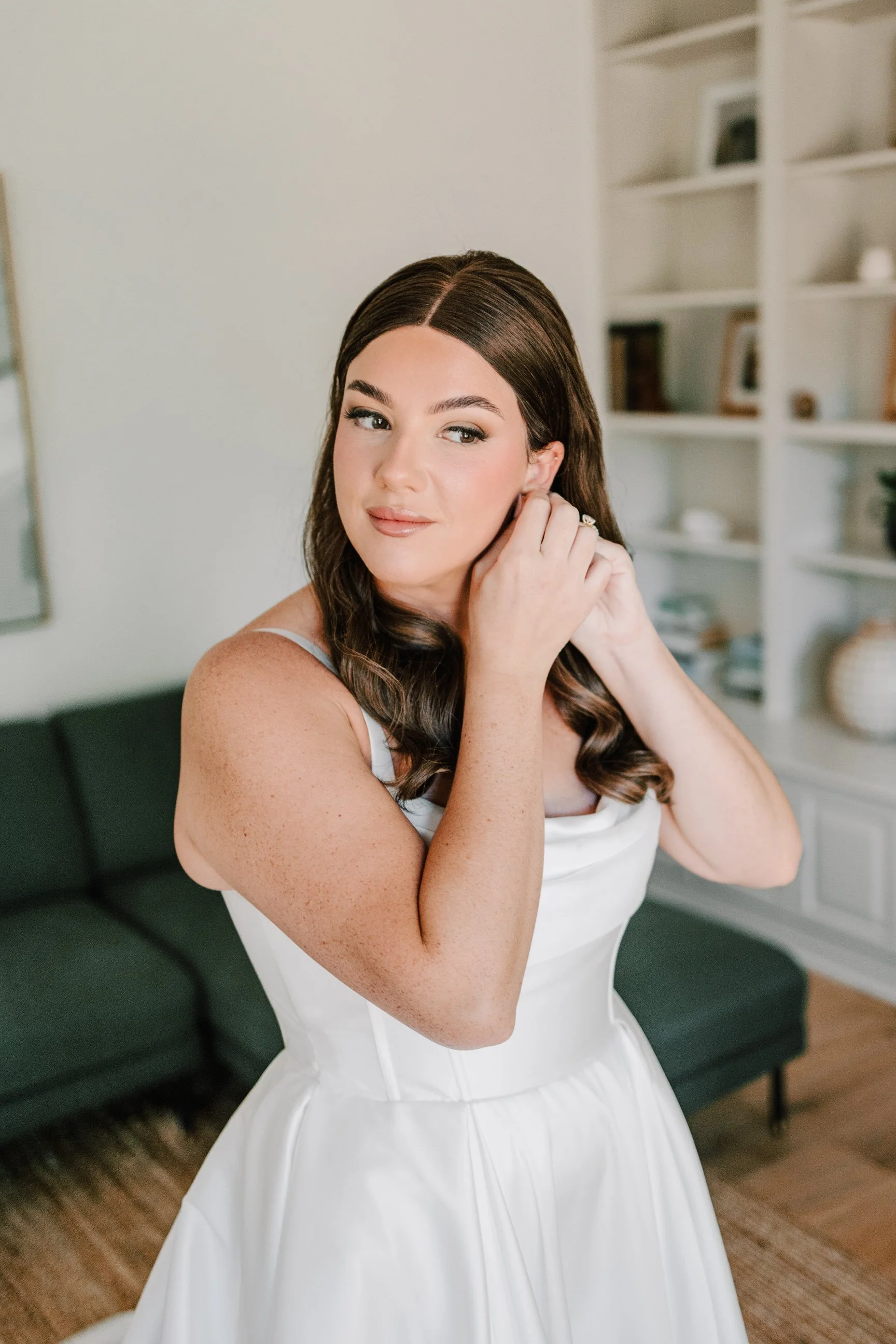 A young woman in a white dress putting on earrings in a living room.