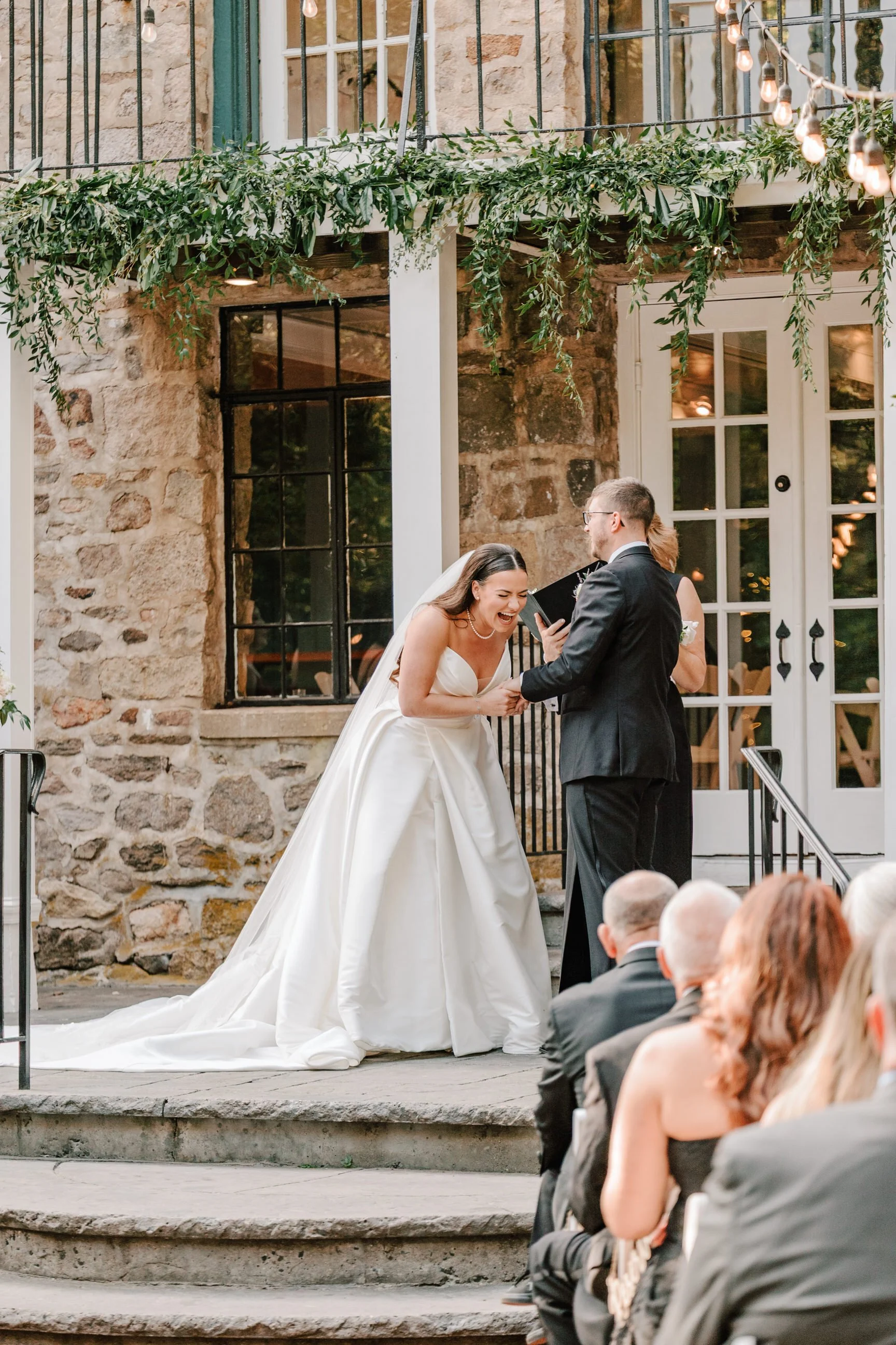 Bride and groom exchange vows during wedding ceremony outside brick building with string lights and greenery