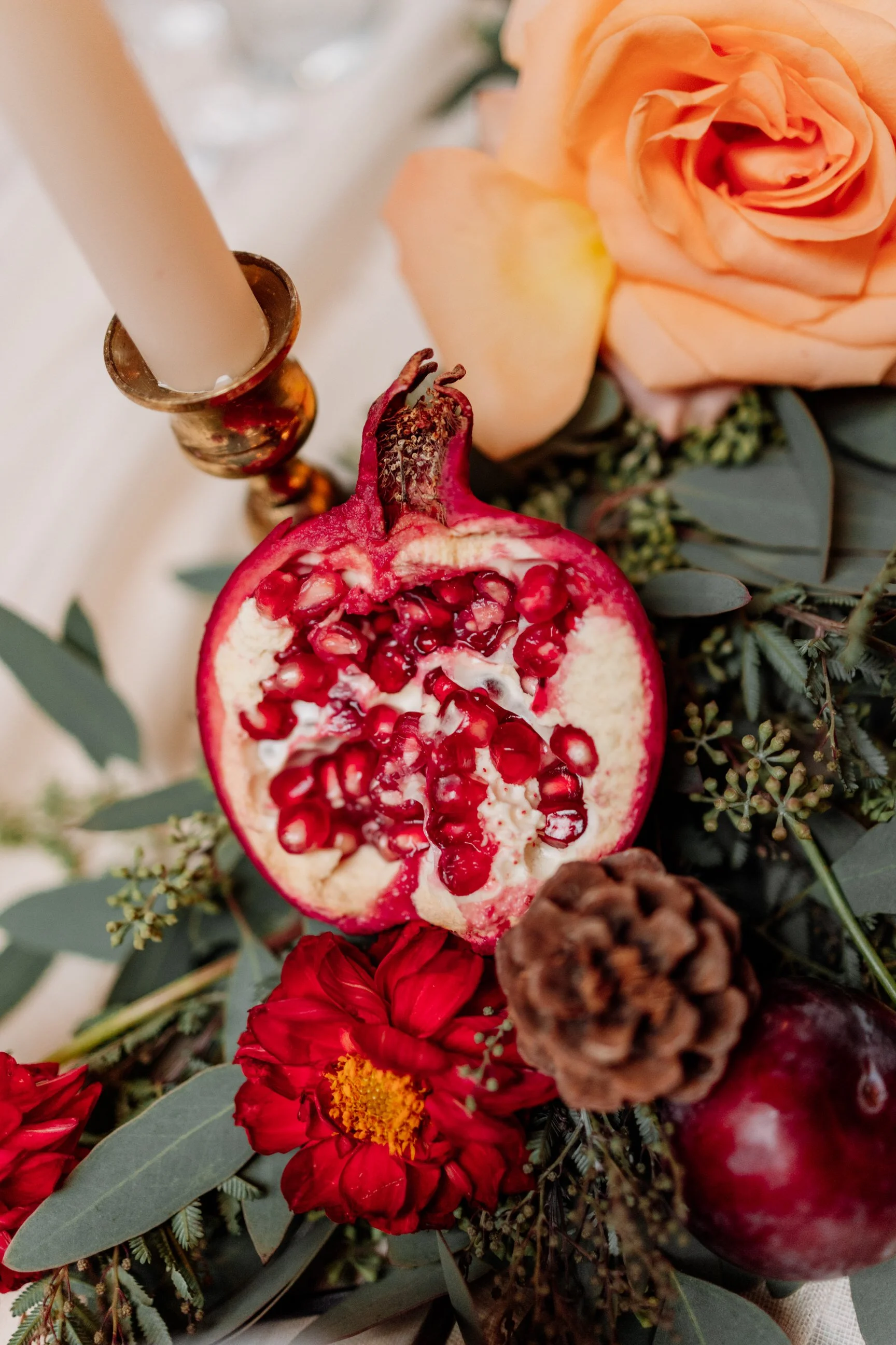 Close-up of a pomegranate half surrounded by flowers, greenery, a candle in a brass holder, and a pinecone on a table.