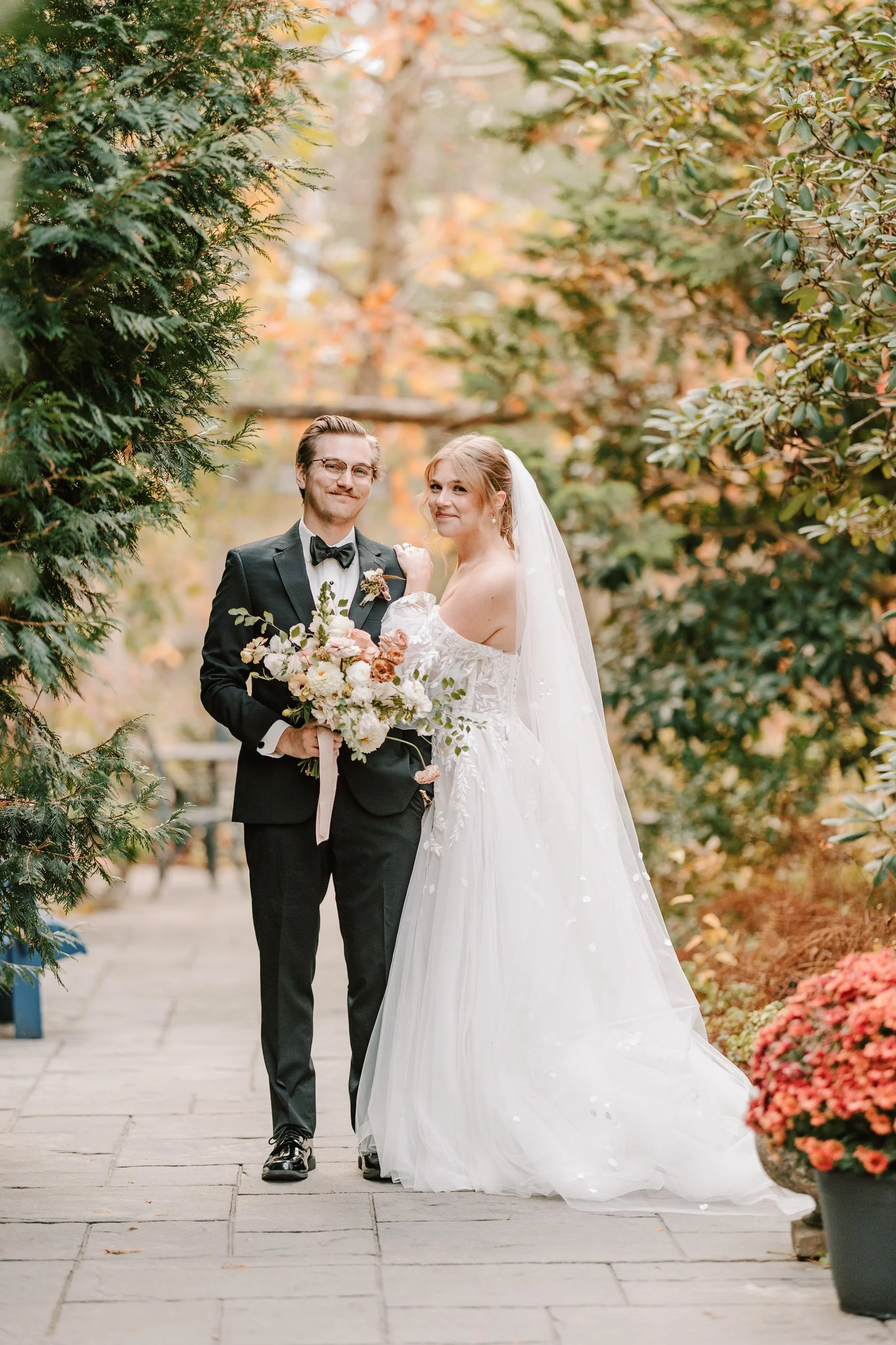 A bride and groom pose in an outdoor garden with autumn foliage. The groom is dressed in a black tuxedo and the bride in a white wedding gown with a veil. The groom holds a bouquet of flowers, and they stand close, smiling. Holly Hedge Estate 