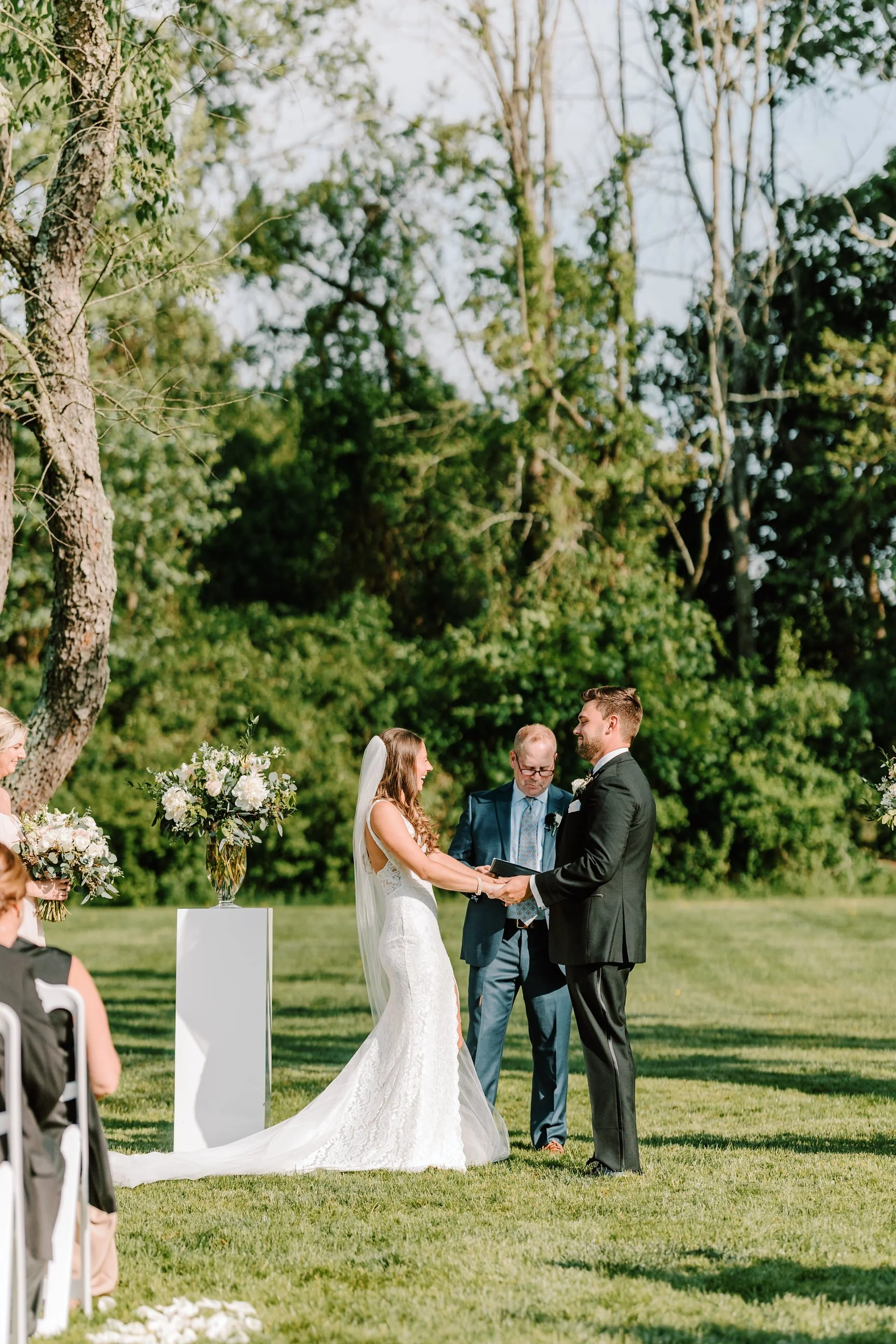 A couple is getting married outdoors, holding hands and exchanging vows, with an officiant standing between them. The bride is in a white gown with a long train and veil, and the groom is in a black suit. Bridesmaids are seated on the left, holding b
