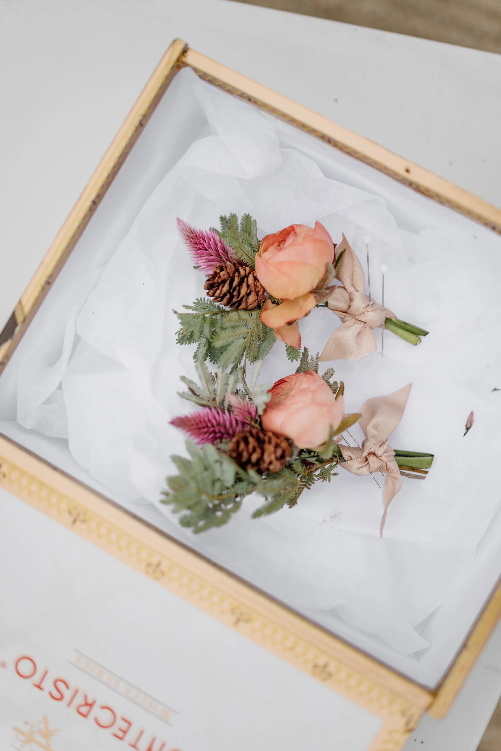 Two boutonnières with peach roses, pink flowers, pinecones, and greenery, lying inside a gift box lined with tissue paper.