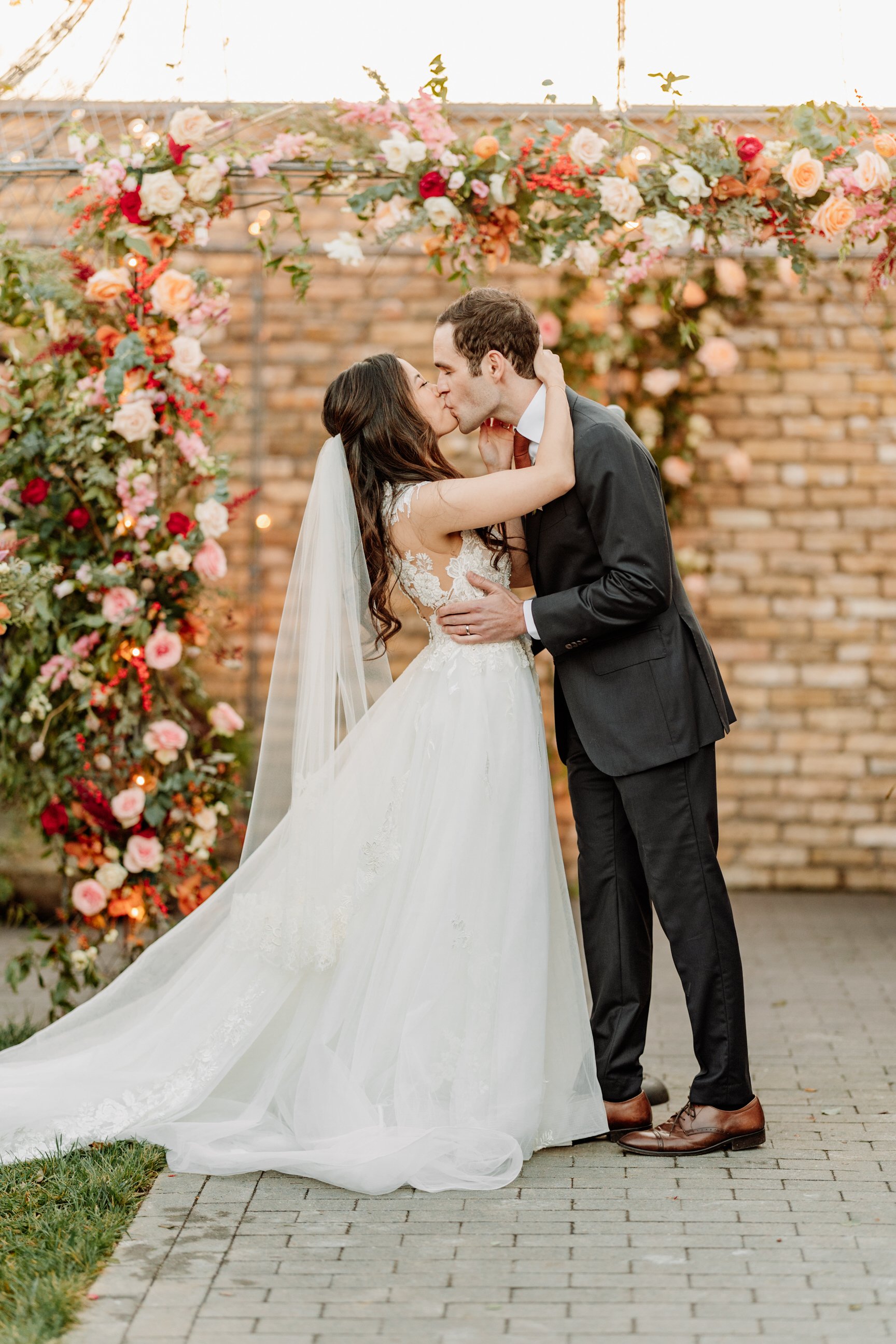 A bride and groom share a kiss during their wedding ceremony under a floral arch.
