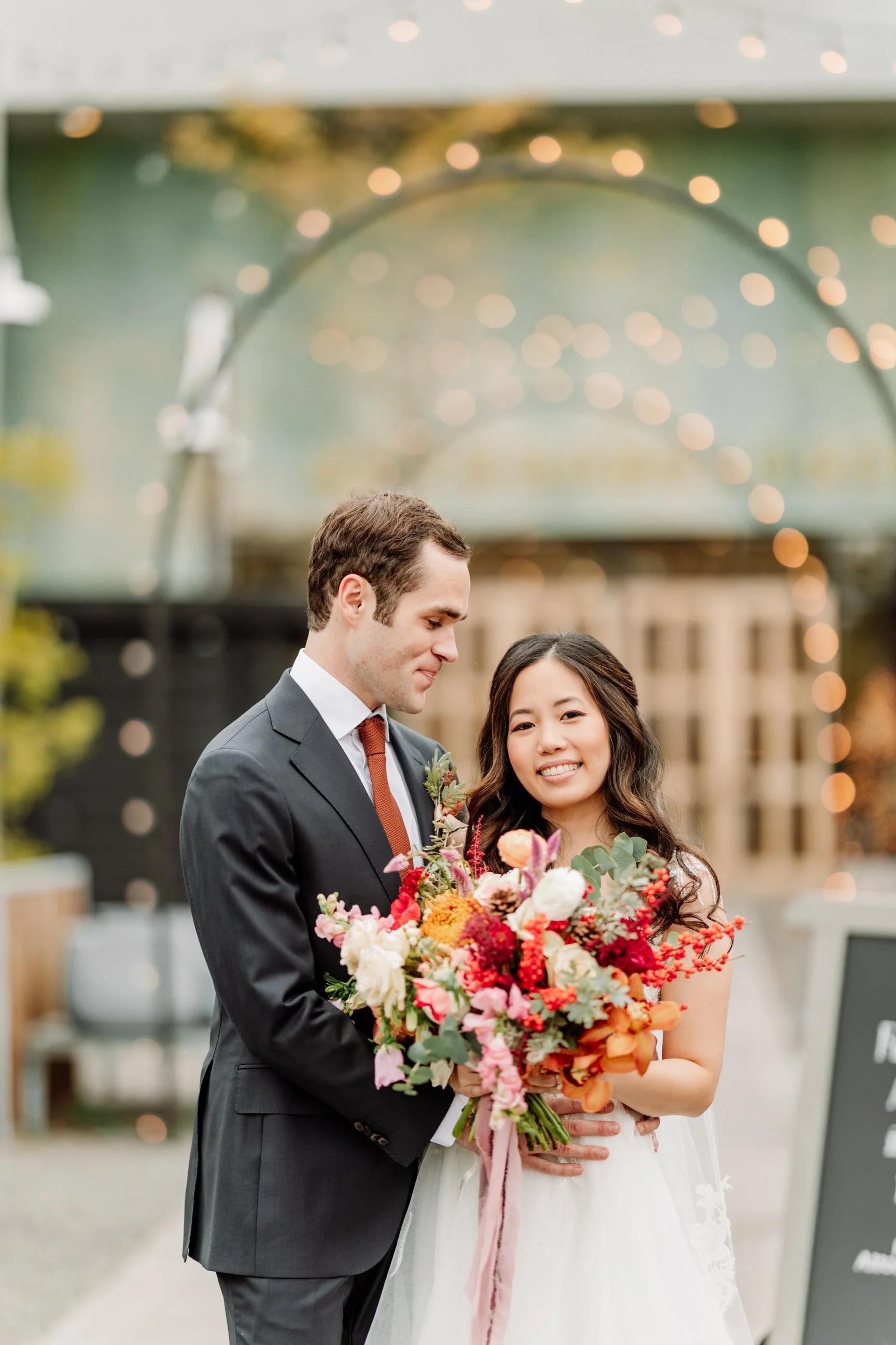 A newlywed couple with the groom in a dark suit and tie and the bride in a white wedding dress, holding a large colorful bouquet of flowers, smiling at a wedding venue decorated with string lights.