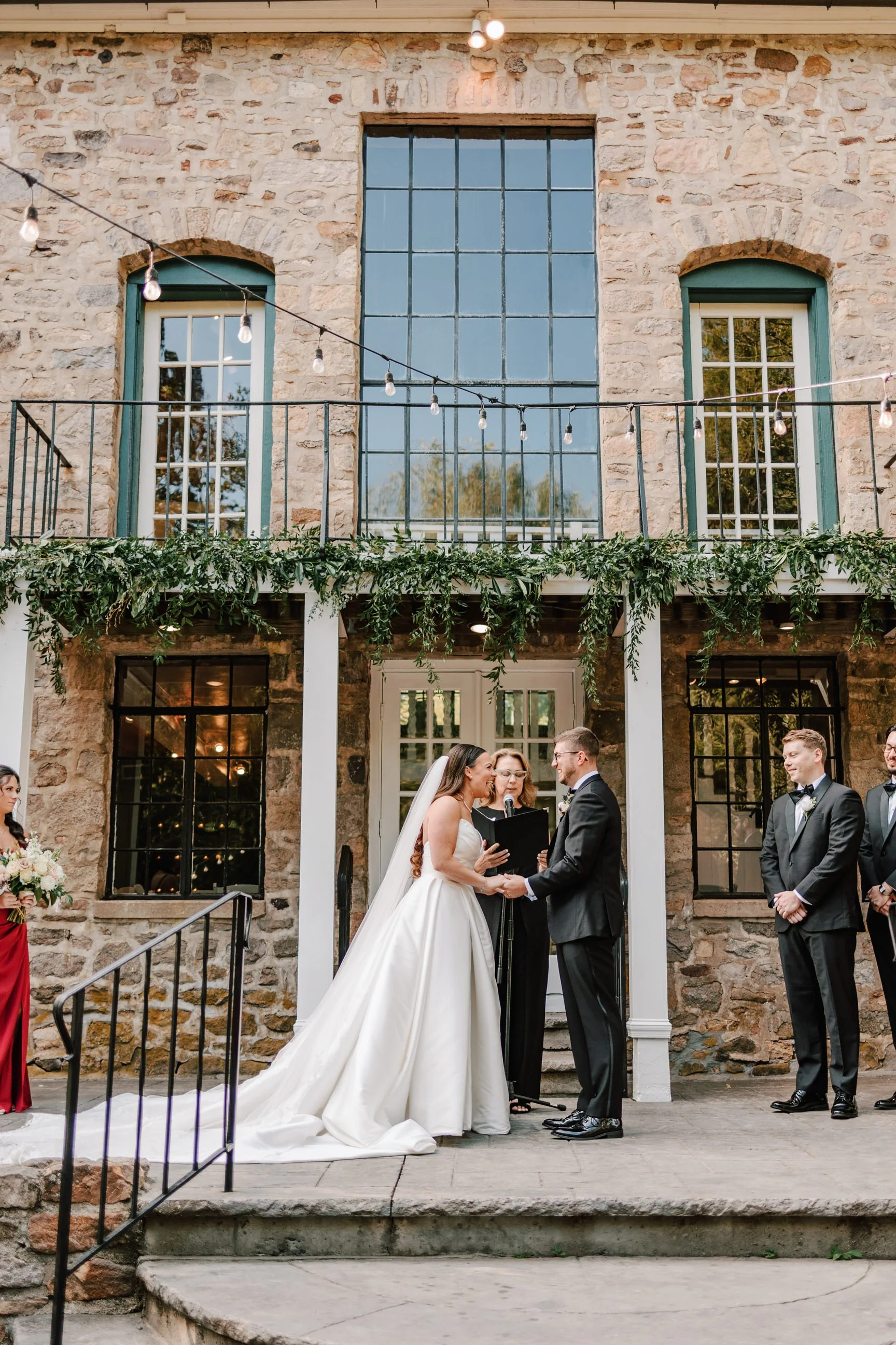 A wedding ceremony with a bride and groom holding hands and exchanging vows in front of an officiant, outside a stone building with large windows, fairy lights, and greenery.