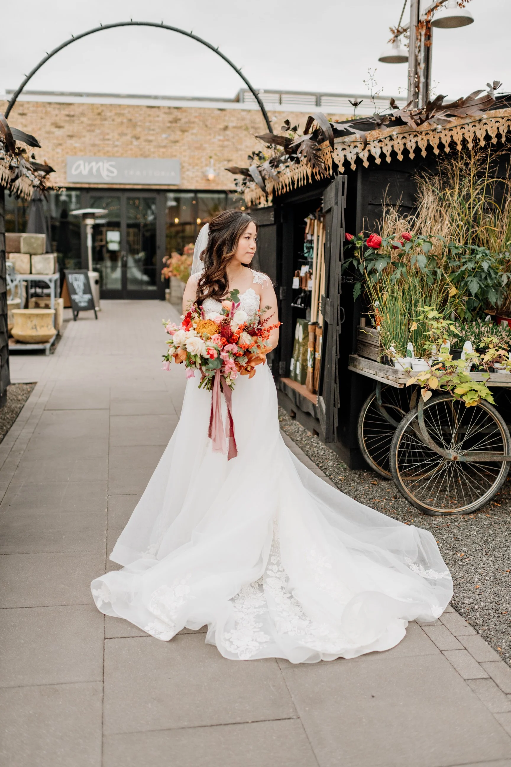 A bride in a white wedding gown holding a bouquet of colorful flowers stands outdoors near a rustic flower stand. She is looking to her right on a cloudy day.