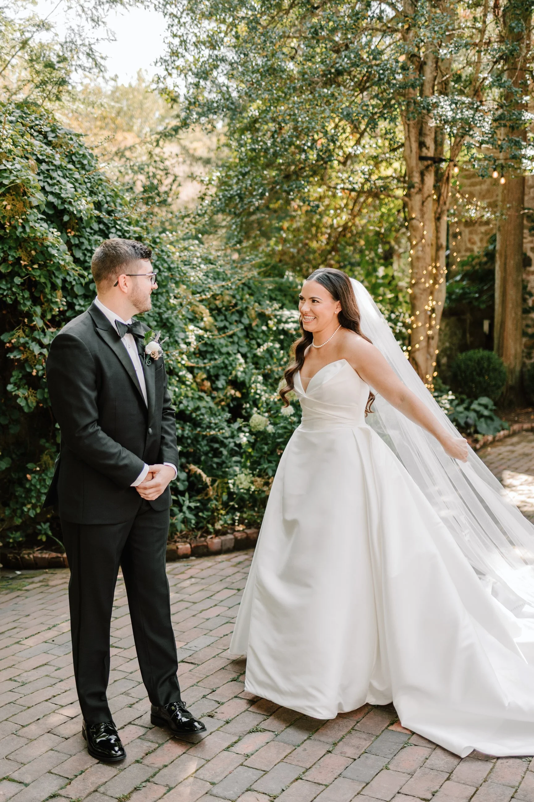 A bride in a white wedding gown and a groom in a black tuxedo stand outdoors on a brick path, smiling at each other, with trees and string lights in the background.