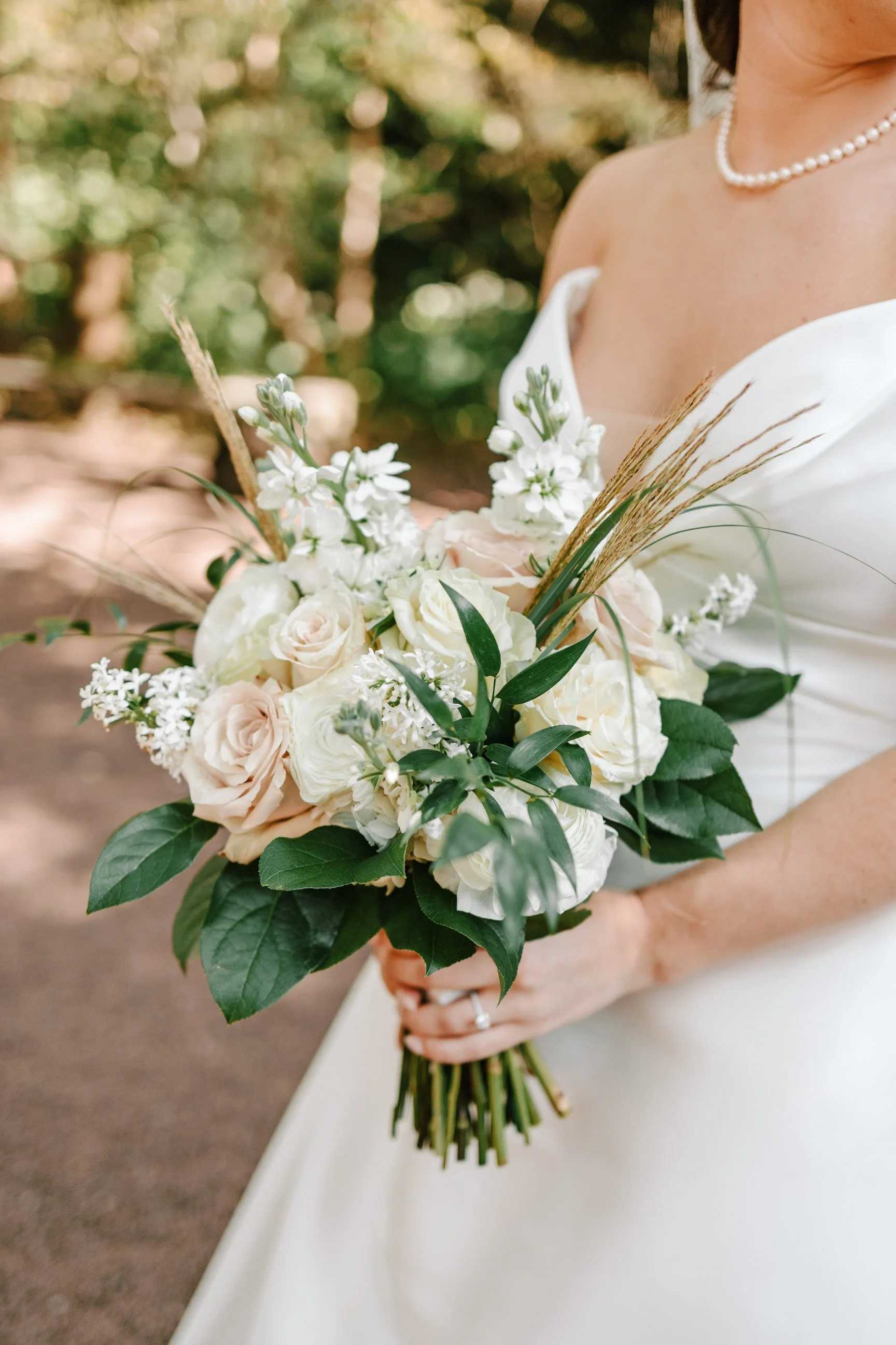Bride holding a bouquet of white and blush roses, white stock flowers, greenery, and wheat stalks, wearing a white off-shoulder dress and pearl necklace.