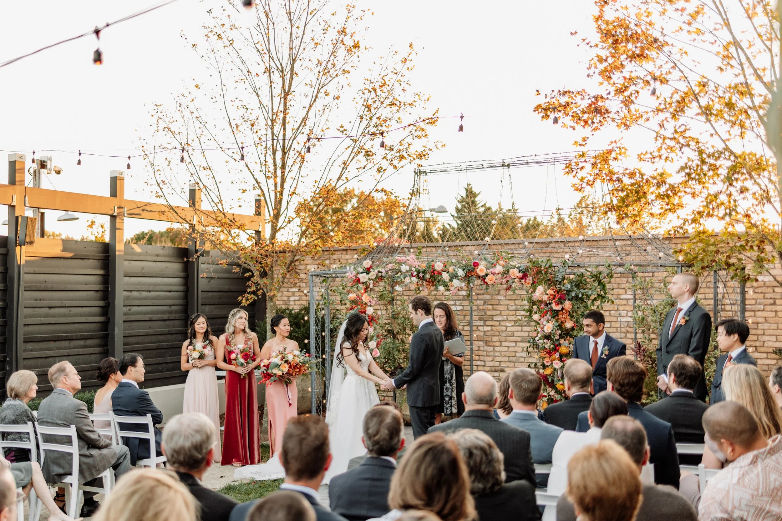Outdoor wedding ceremony with bride and groom holding hands, officiant nearby, surrounded by guests, decorated with floral arch and trees with autumn leaves
