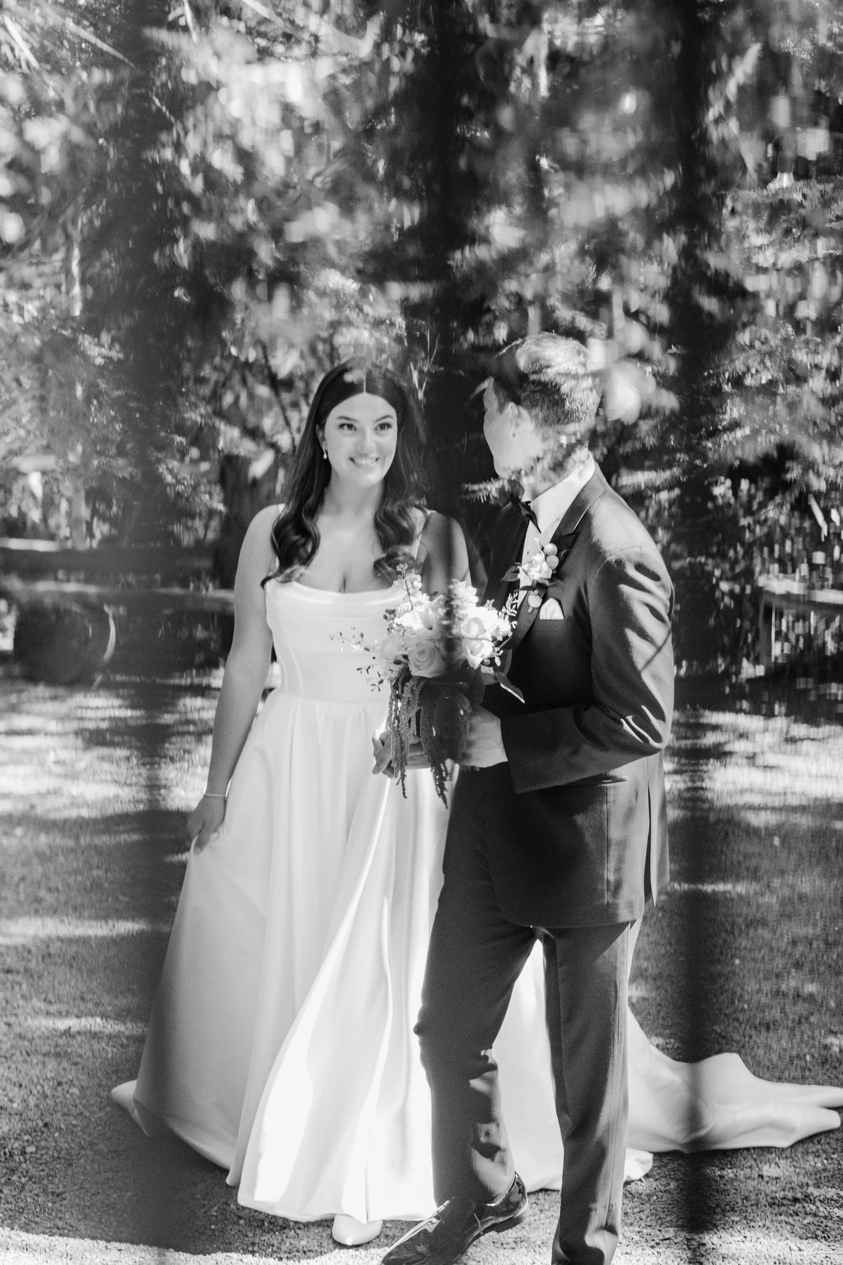 Black and white photo of a bride and groom standing outdoors in a wooded area, smiling and holding a bouquet of flowers, with sunlight filtering through trees.