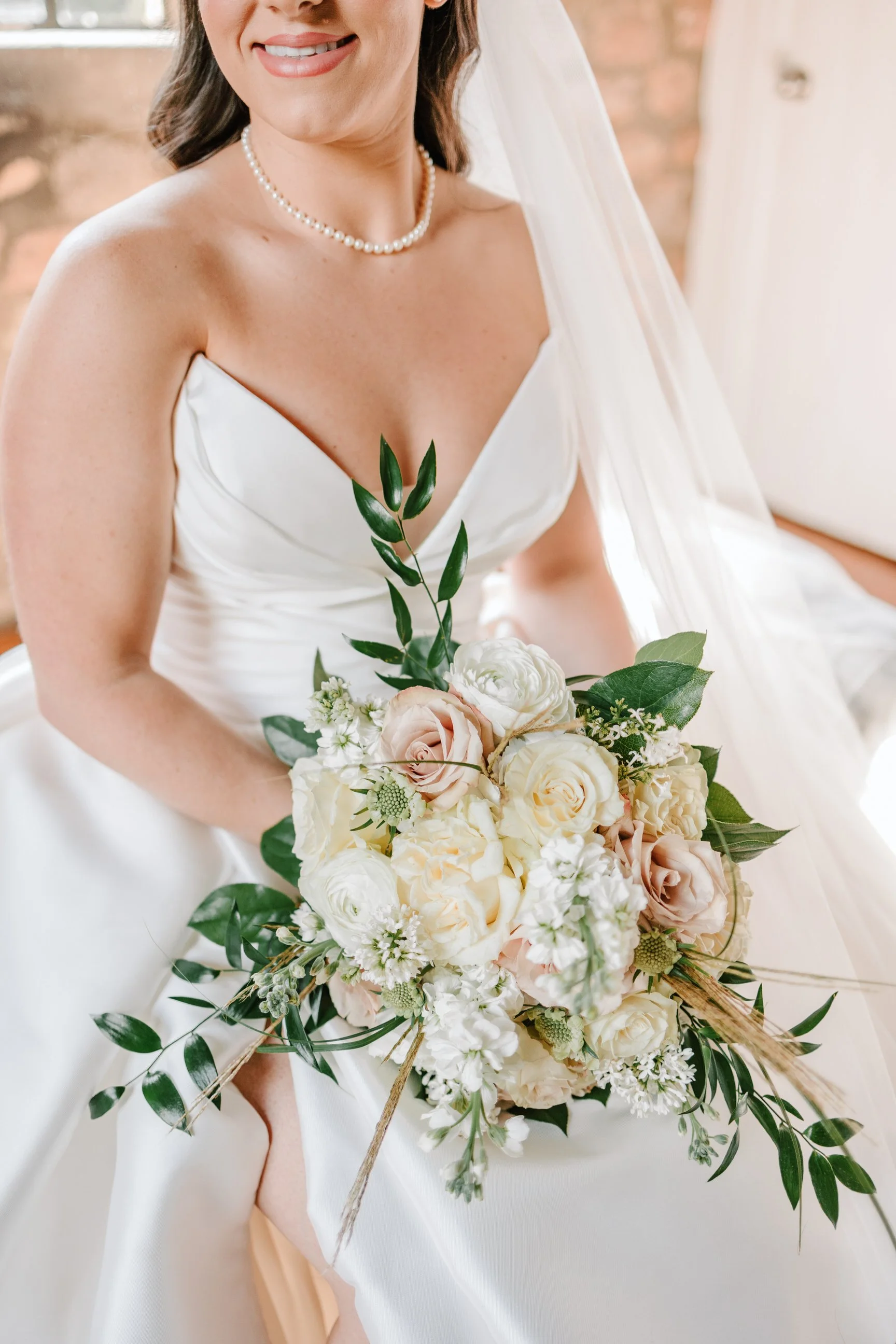 A bride in a white wedding dress holding a bouquet of white and pale pink roses with green foliage.
