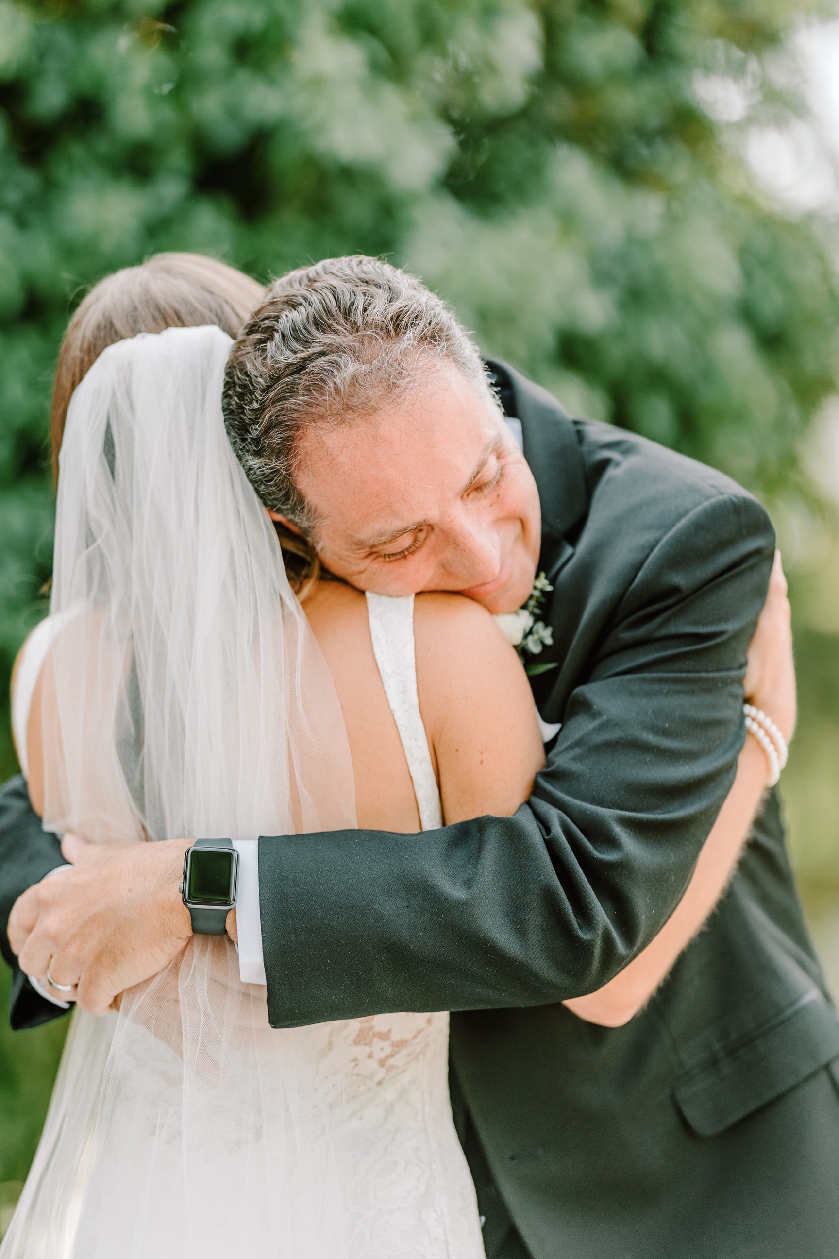 A bride and groom hugging outdoors, with the groom smiling and the bride wearing a veil and white dress.