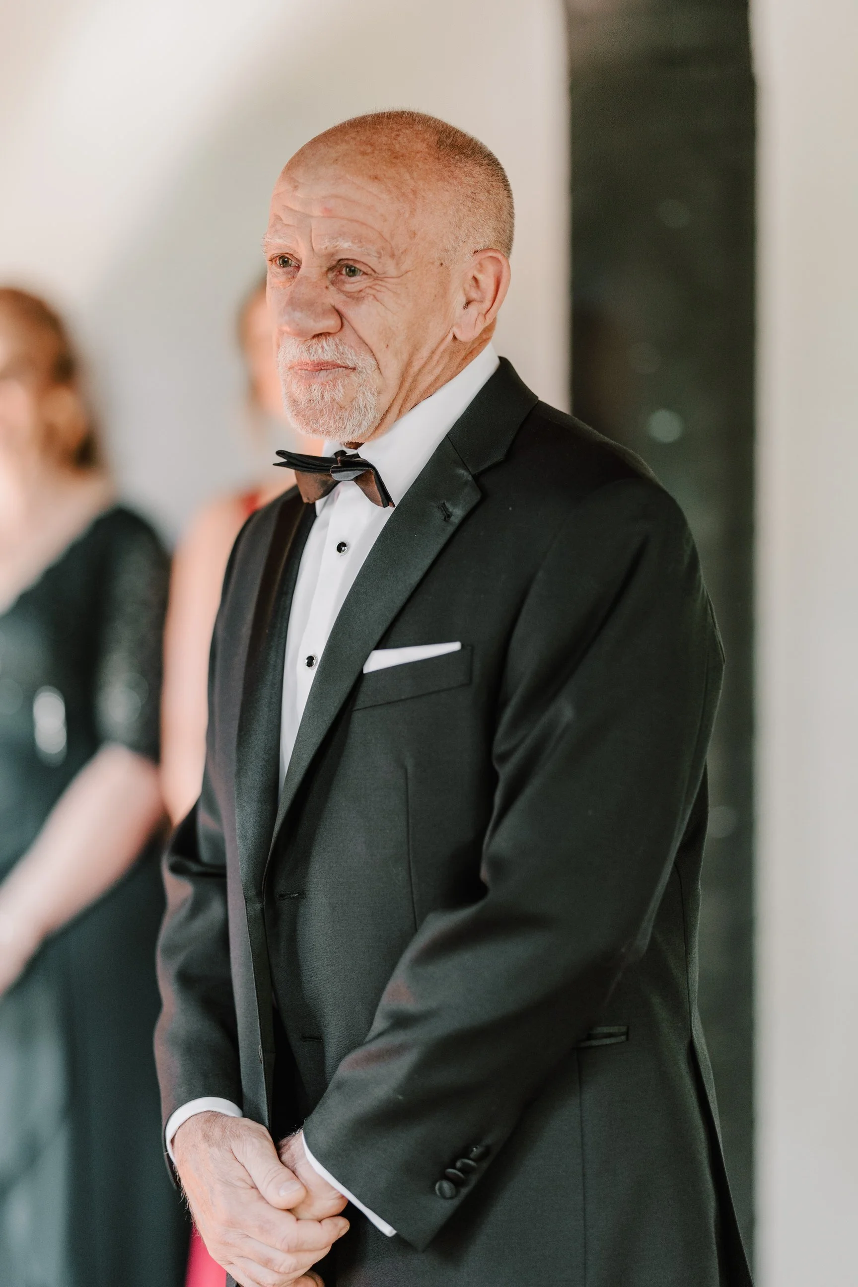 An elderly man in a black tuxedo with a bow tie and white dress shirt, standing with his hands clasped, at what appears to be a formal event.