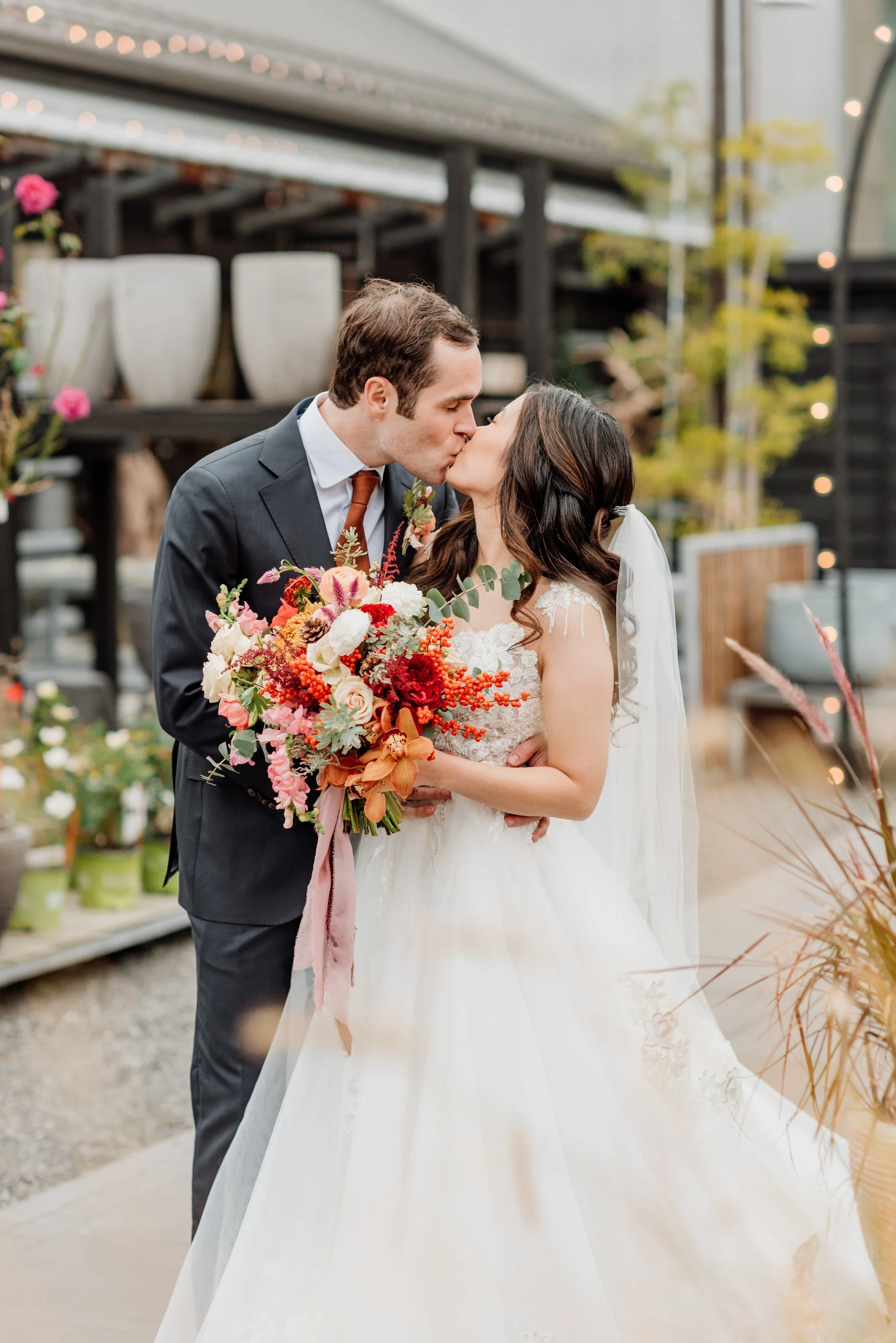 A bride and groom kiss at their wedding, with the bride holding a large colorful bouquet of flowers, in a decorated outdoor setting.
