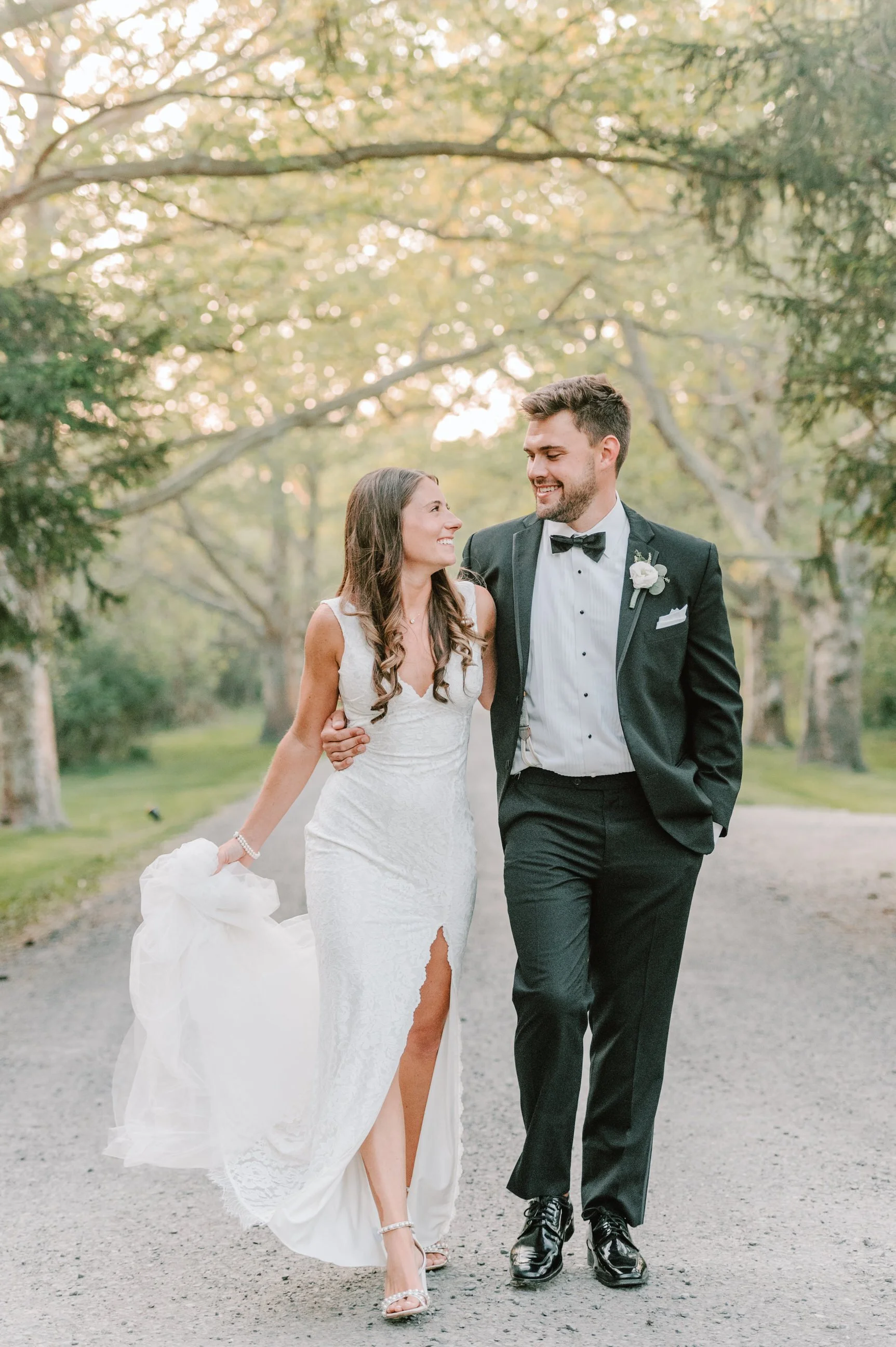 A newlywed couple walking down a tree-lined path, smiling and looking at each other, dressed in wedding attire with the bride in a white lace gown and the groom in a black tuxedo, outdoor setting with sunlight filtering through trees.