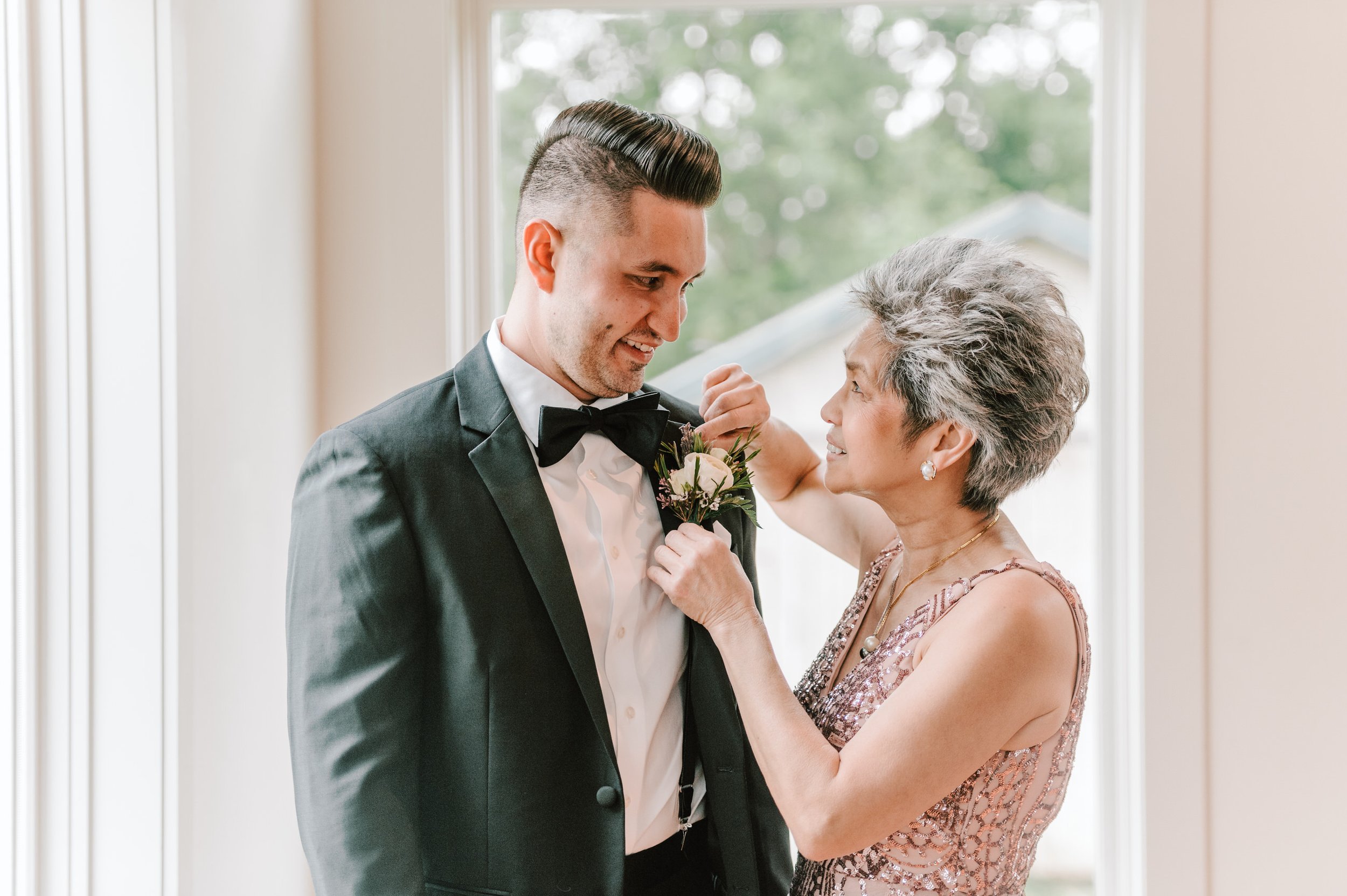 A young man in a black tuxedo and bow tie is being boutonniere pinned by an older woman in a pink, sequined dress. They are smiling at each other near a window with green trees outside.