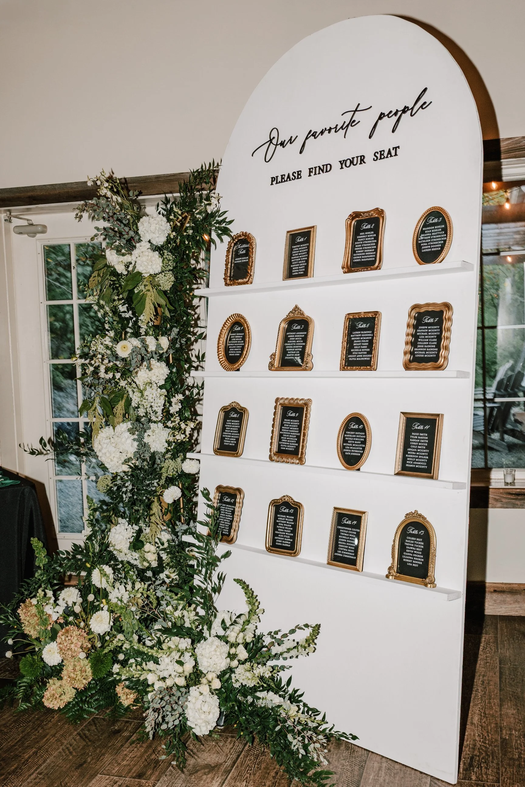 Seating chart display with a white board, black and gold frames, and floral decorations at a wedding or event.