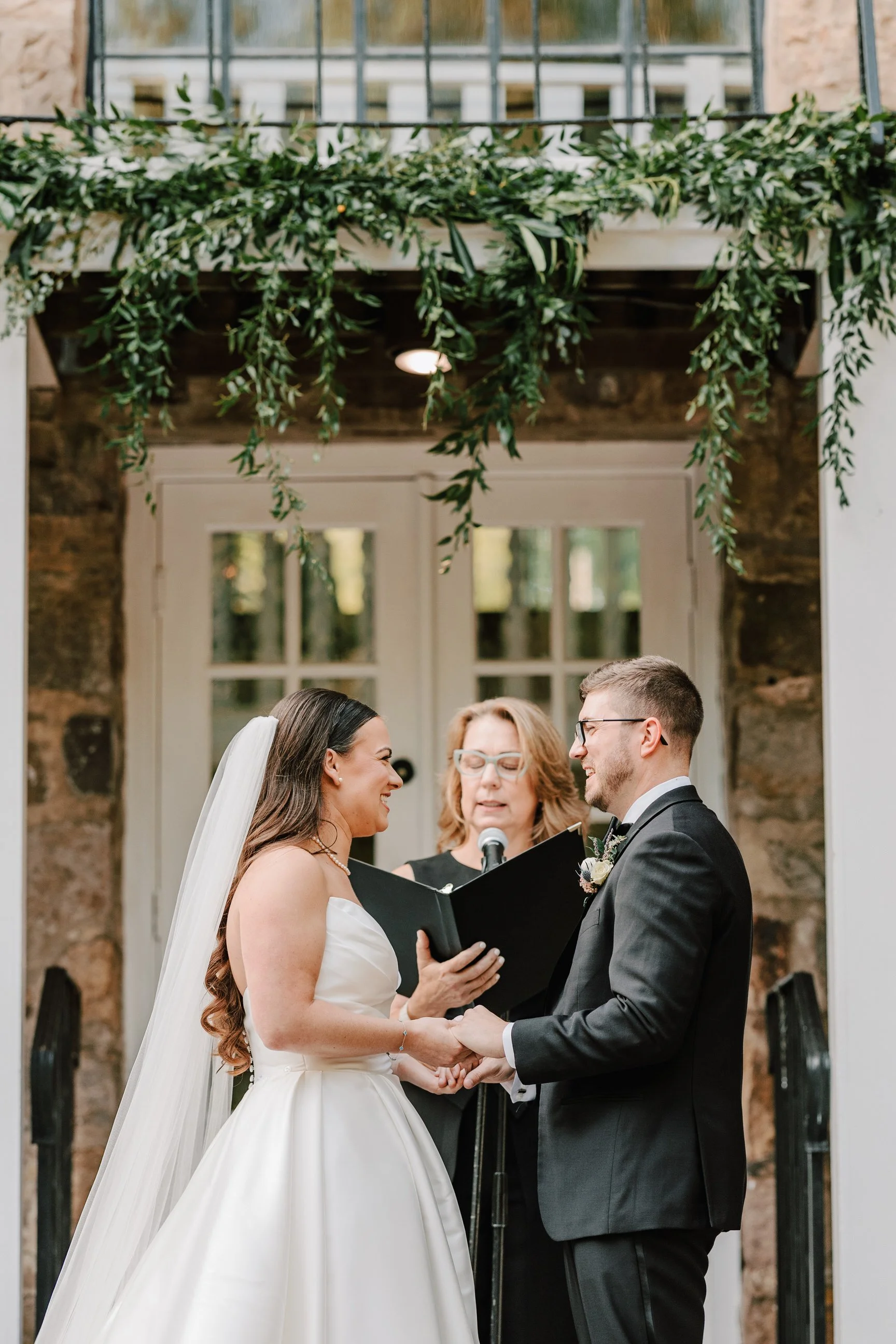 A bride and groom exchanging vows during a wedding ceremony outdoors, with an officiant standing behind them, under greenery and a rustic stone building backdrop.