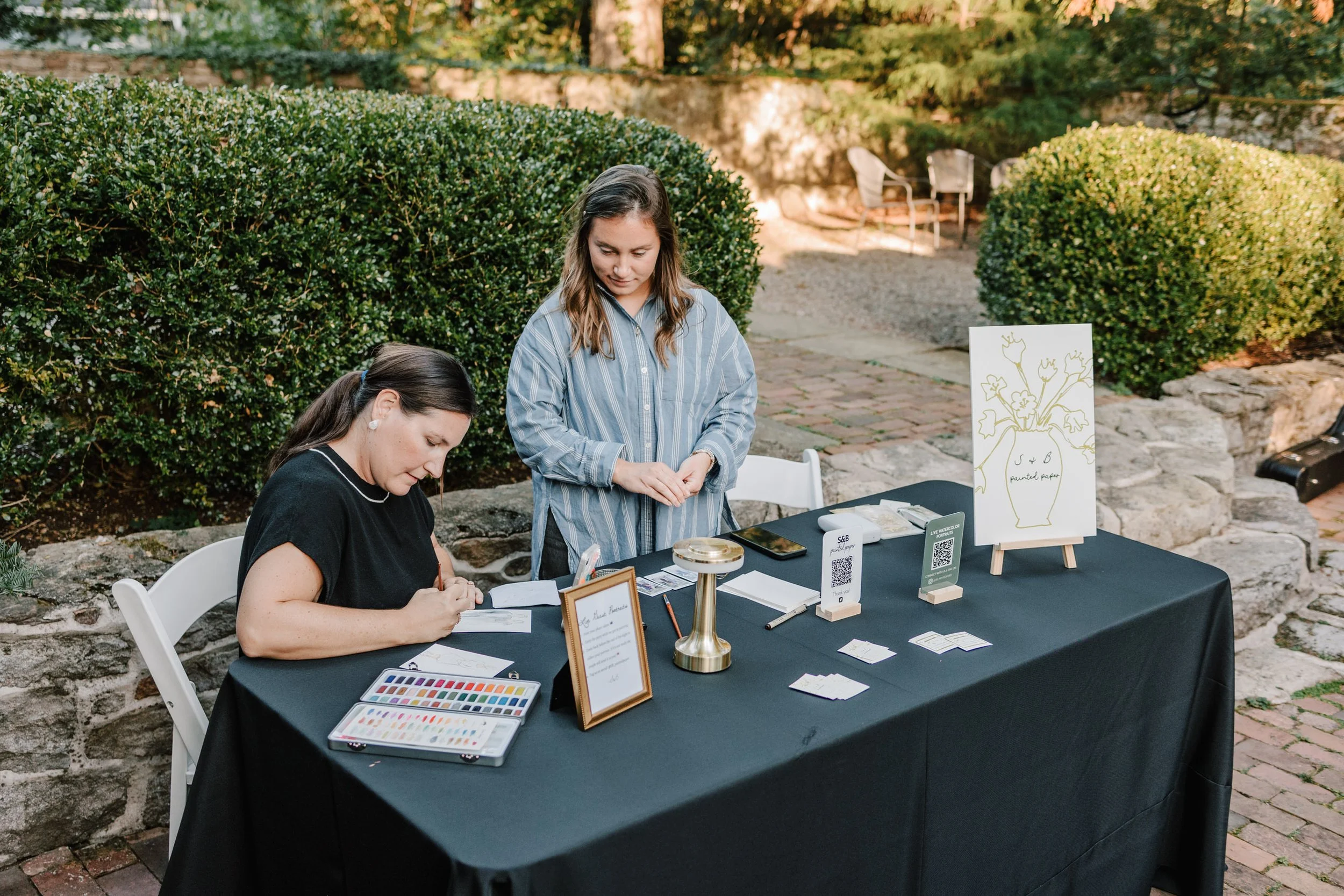 Two women standing at a table with business cards, QR code signs, and artwork in an outdoor setting with bushes and stone walls.