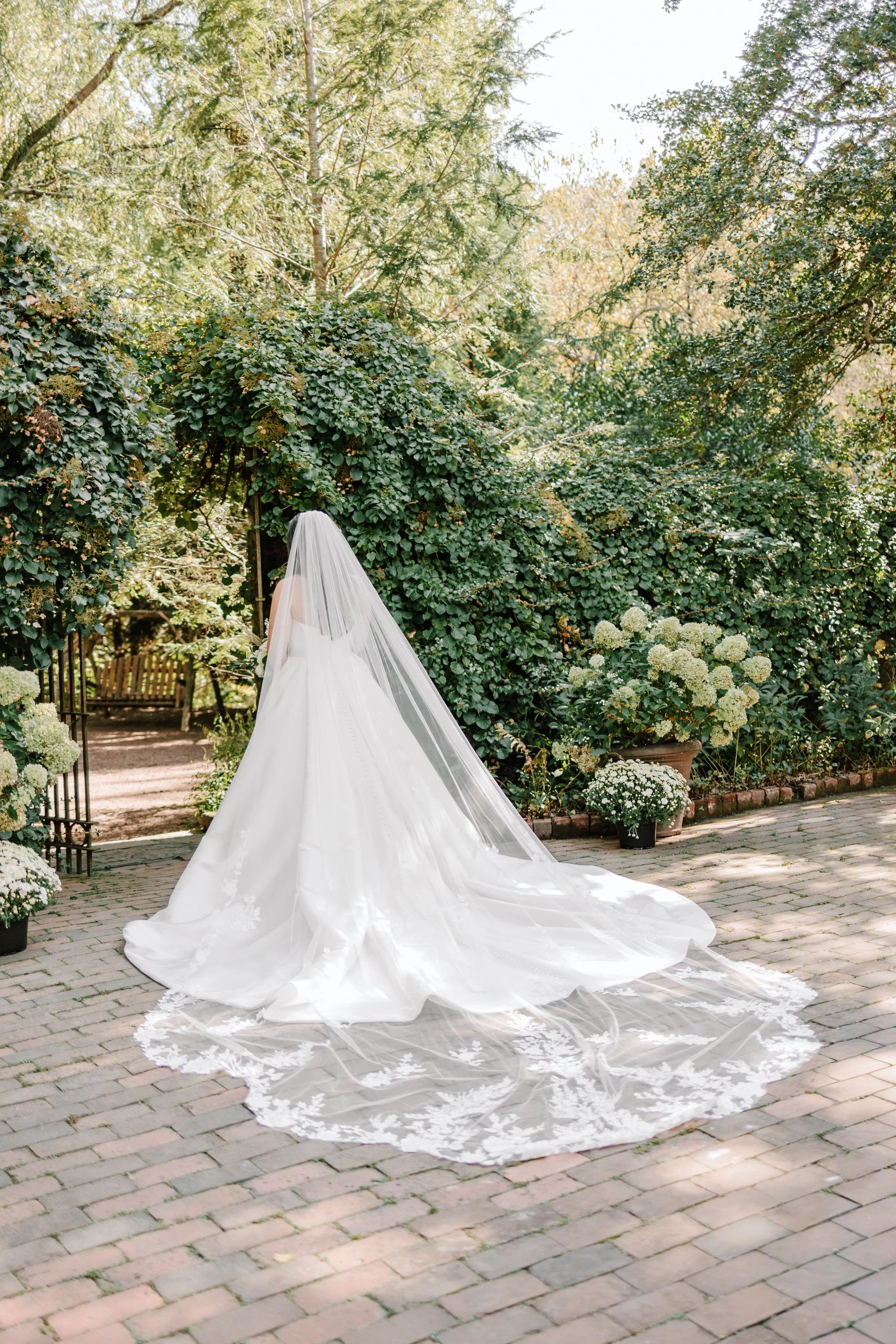 A bride in a white wedding gown with a long train and veil standing outdoors in a garden, surrounded by greenery and white flowers.