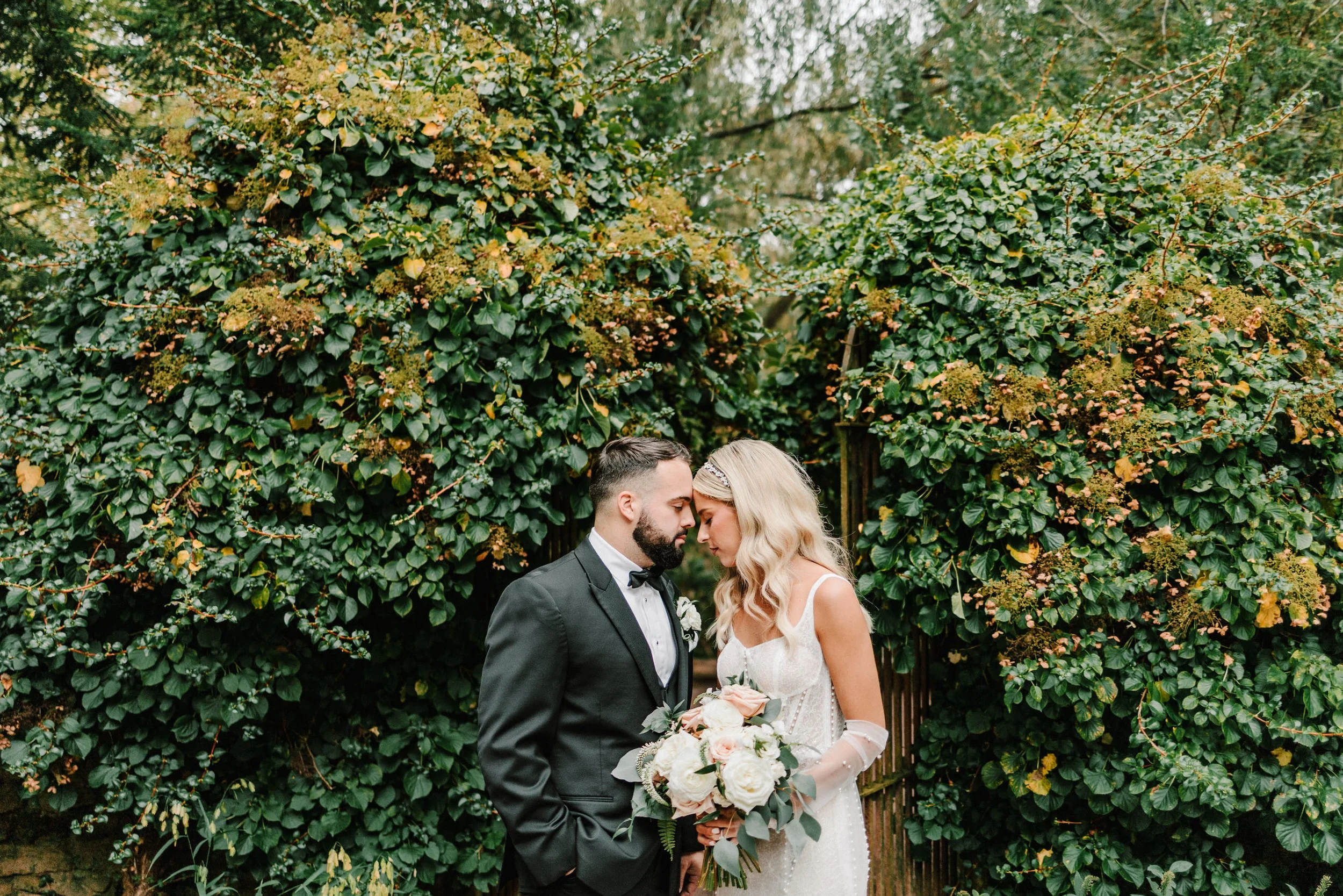 Bride and groom standing closely in front of lush green foliage, the bride holding a bouquet of white and peach flowers.
