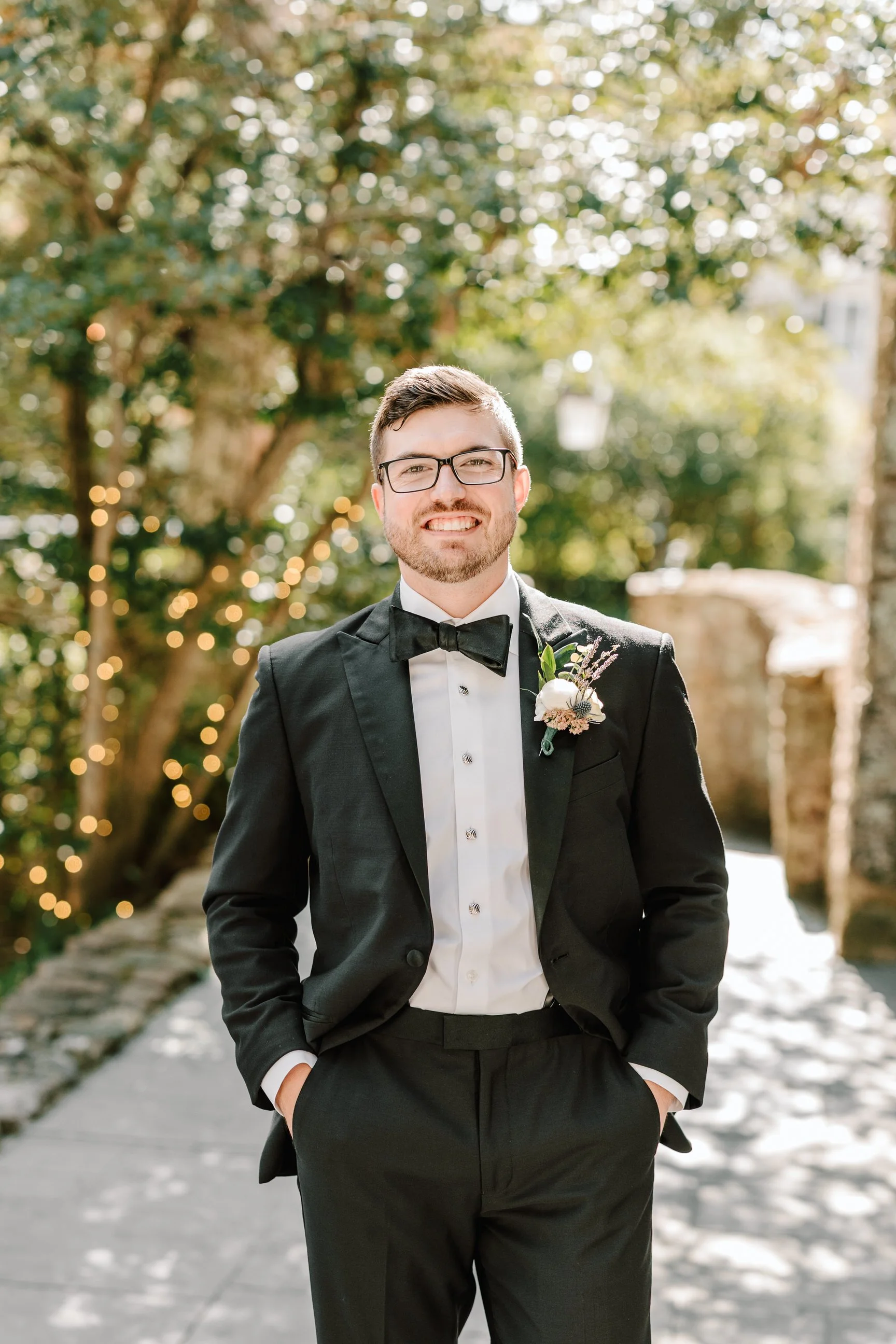 A man in a black tuxedo with a bow tie and glasses, standing outdoors, smiling, with greenery and string lights in the background.