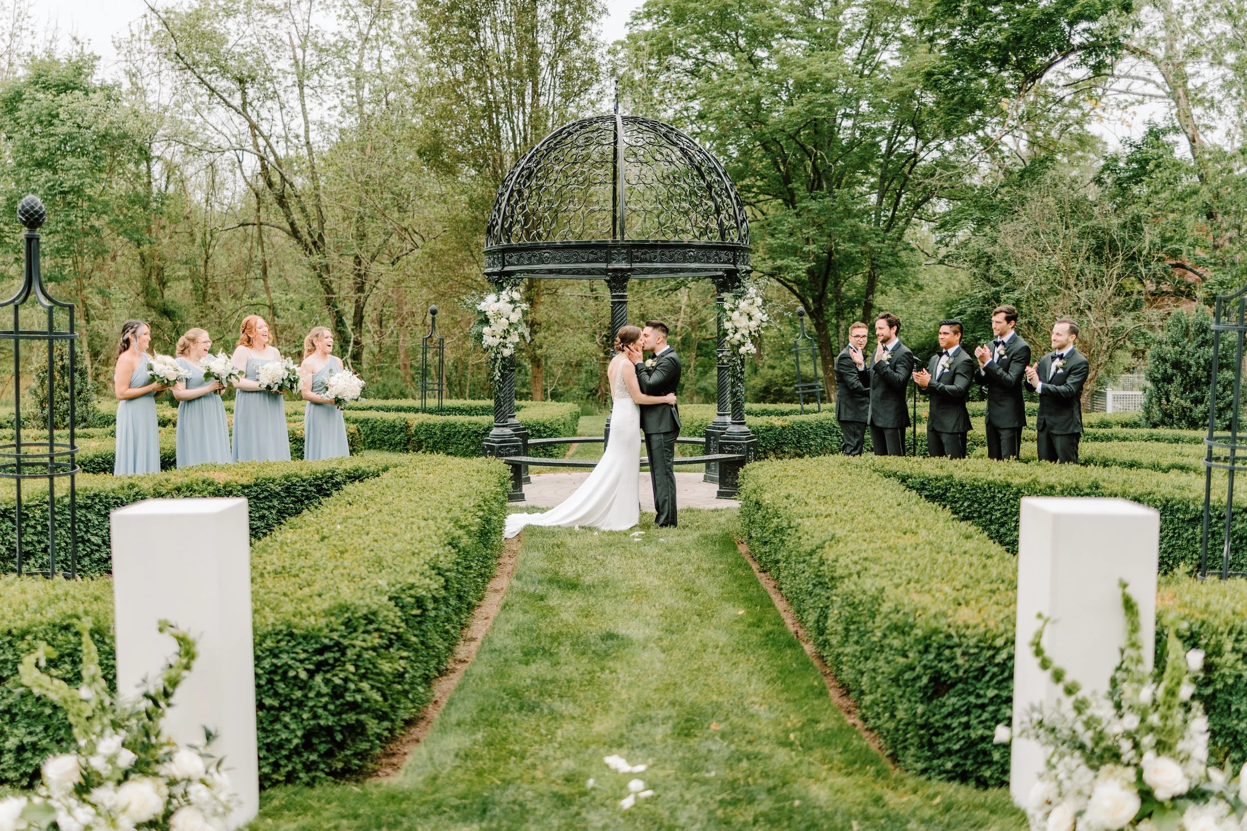 Wedding ceremony outdoors with a bride and groom kissing under a black gazebo, surrounded by bridesmaids in light blue dresses and groomsmen in black suits, in a lush green garden.
