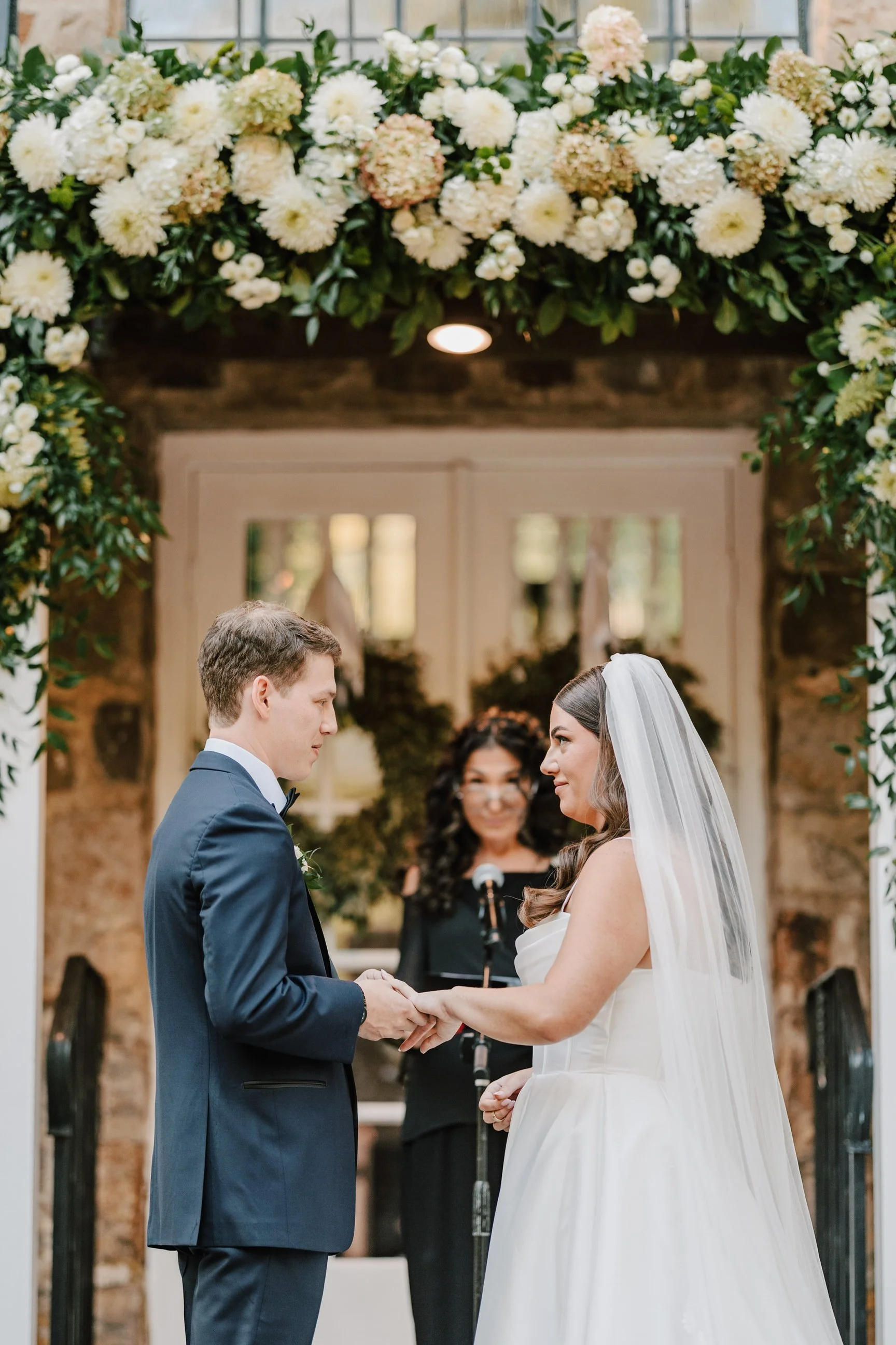 A bride and groom holding hands during their wedding ceremony, with an officiant standing behind them in a floral archway.