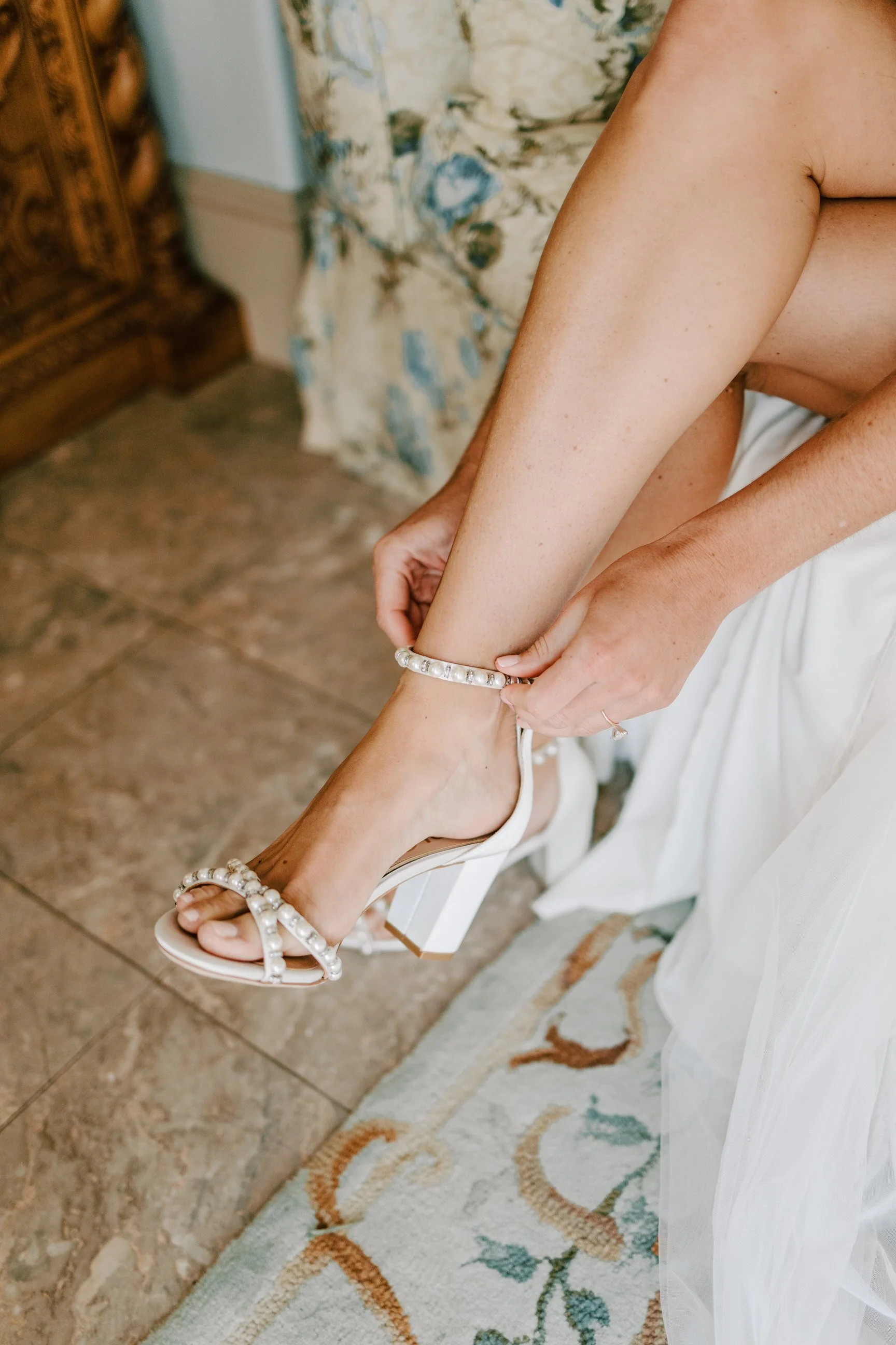 A bride is putting on a white high-heeled sandal with pearl embellishments, fastening the ankle strap.