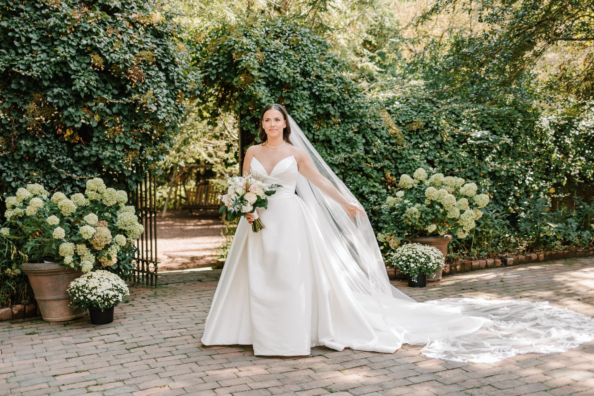 Bride in a white wedding gown holding a bouquet, standing on a brick pathway with greenery and white flowers in the background.