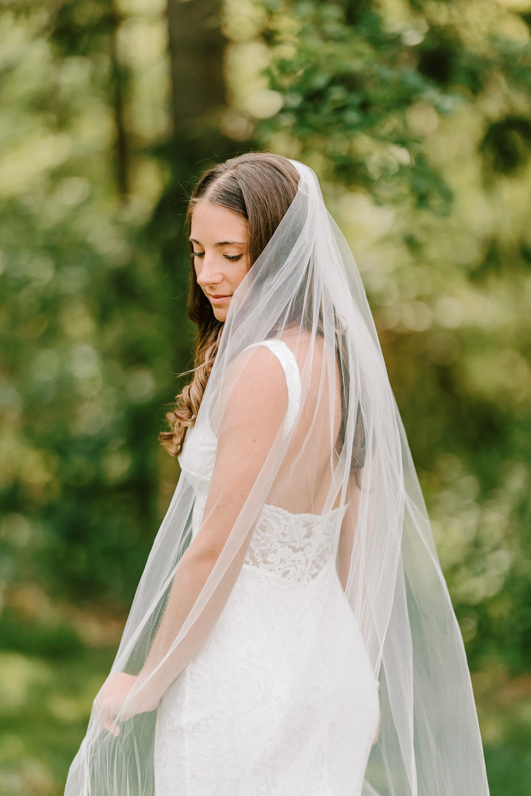 A bride outdoors wearing a white lace wedding dress and a sheer veil, standing with her head slightly bowed, surrounded by greenery.