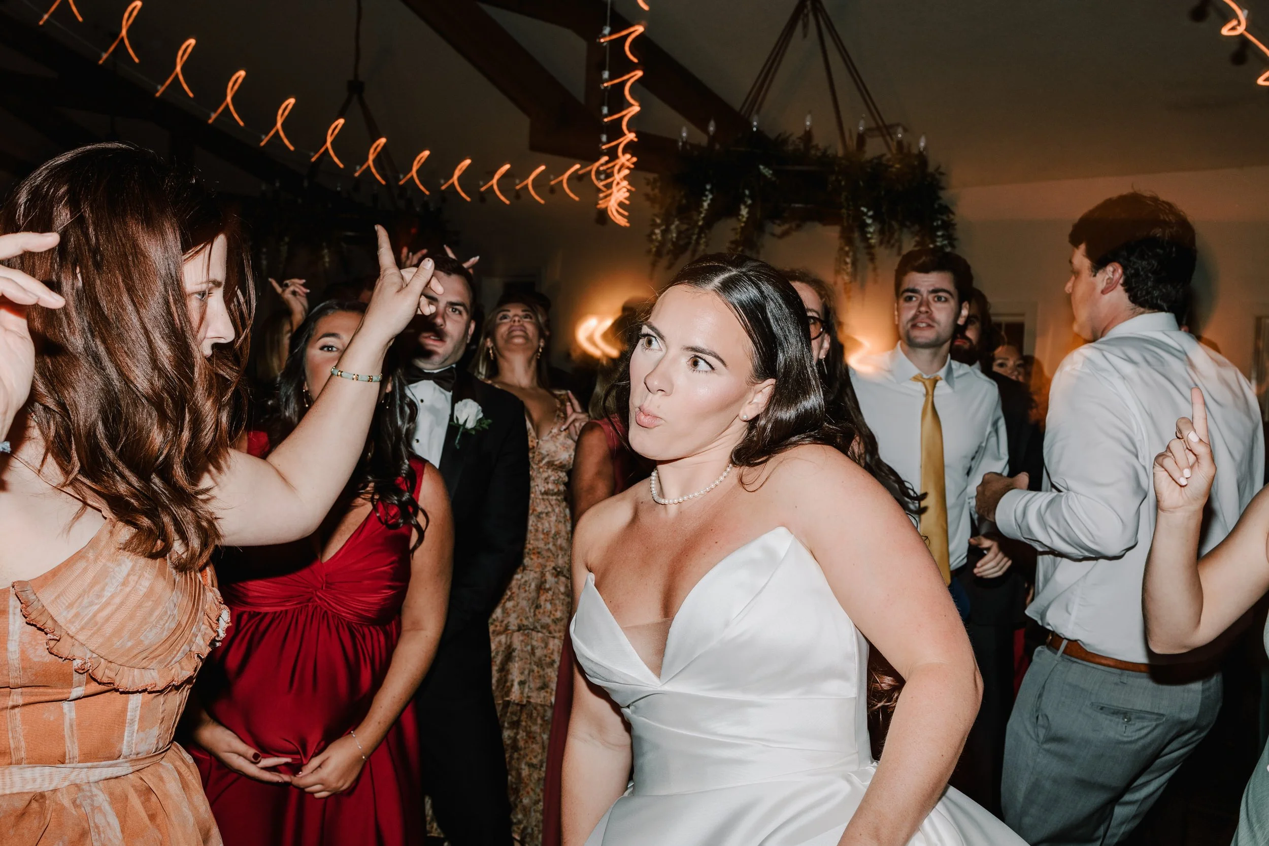 A woman in a white wedding dress making a playful face on the dance floor at a wedding reception, surrounded by friends and family dancing.