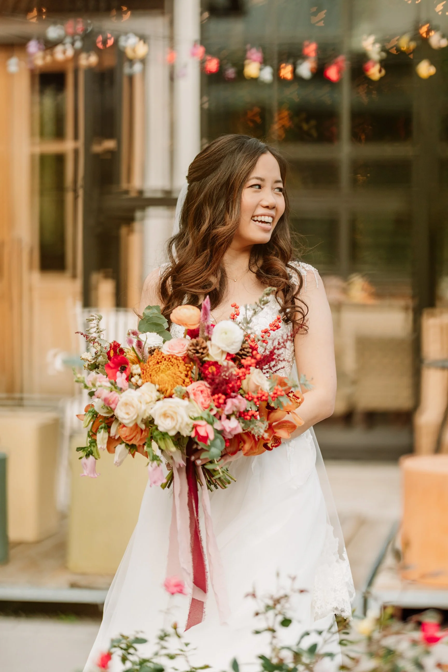 A woman in a wedding dress holding a colorful bouquet of flowers, standing outdoors with blurred background.