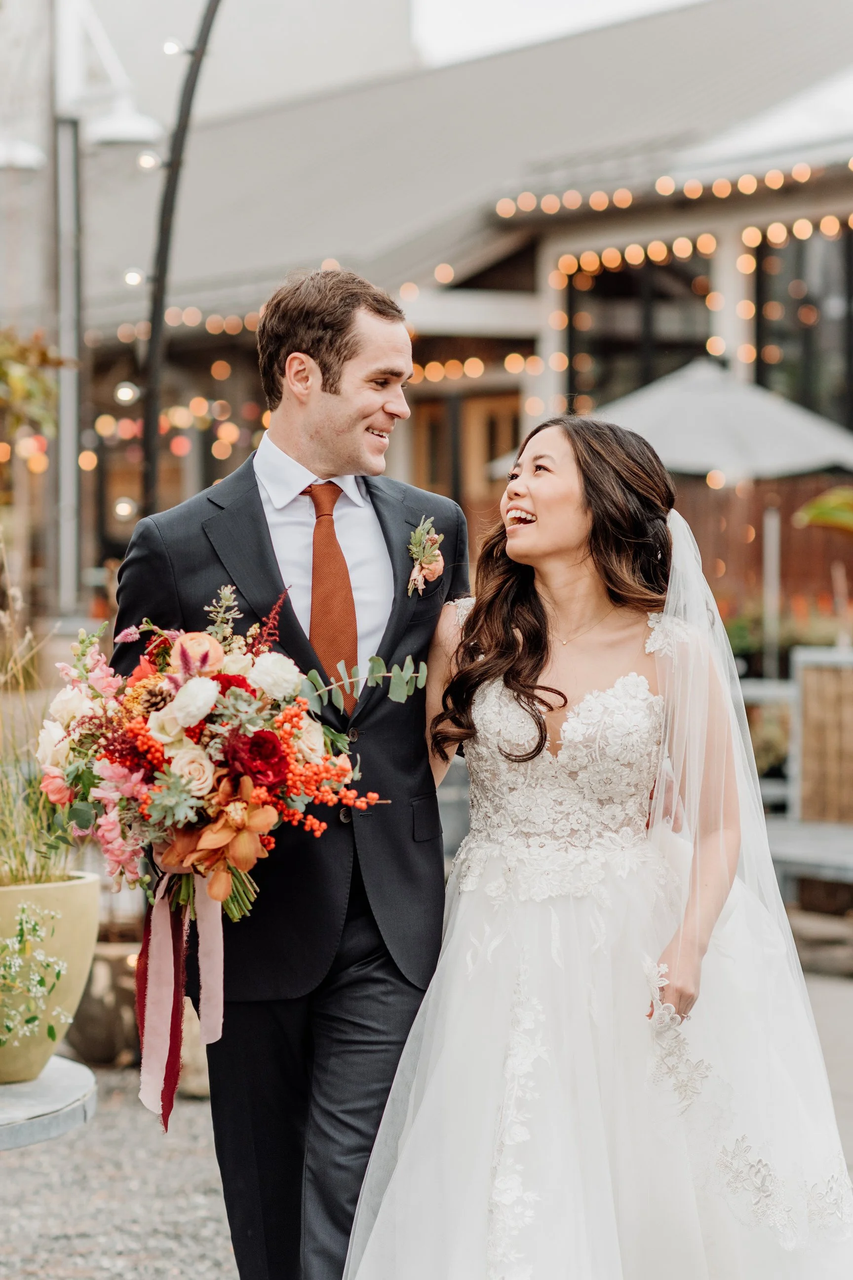 A smiling bride and groom celebrating outdoors with string lights and a building in the background, the groom holding a bouquet of colorful flowers.